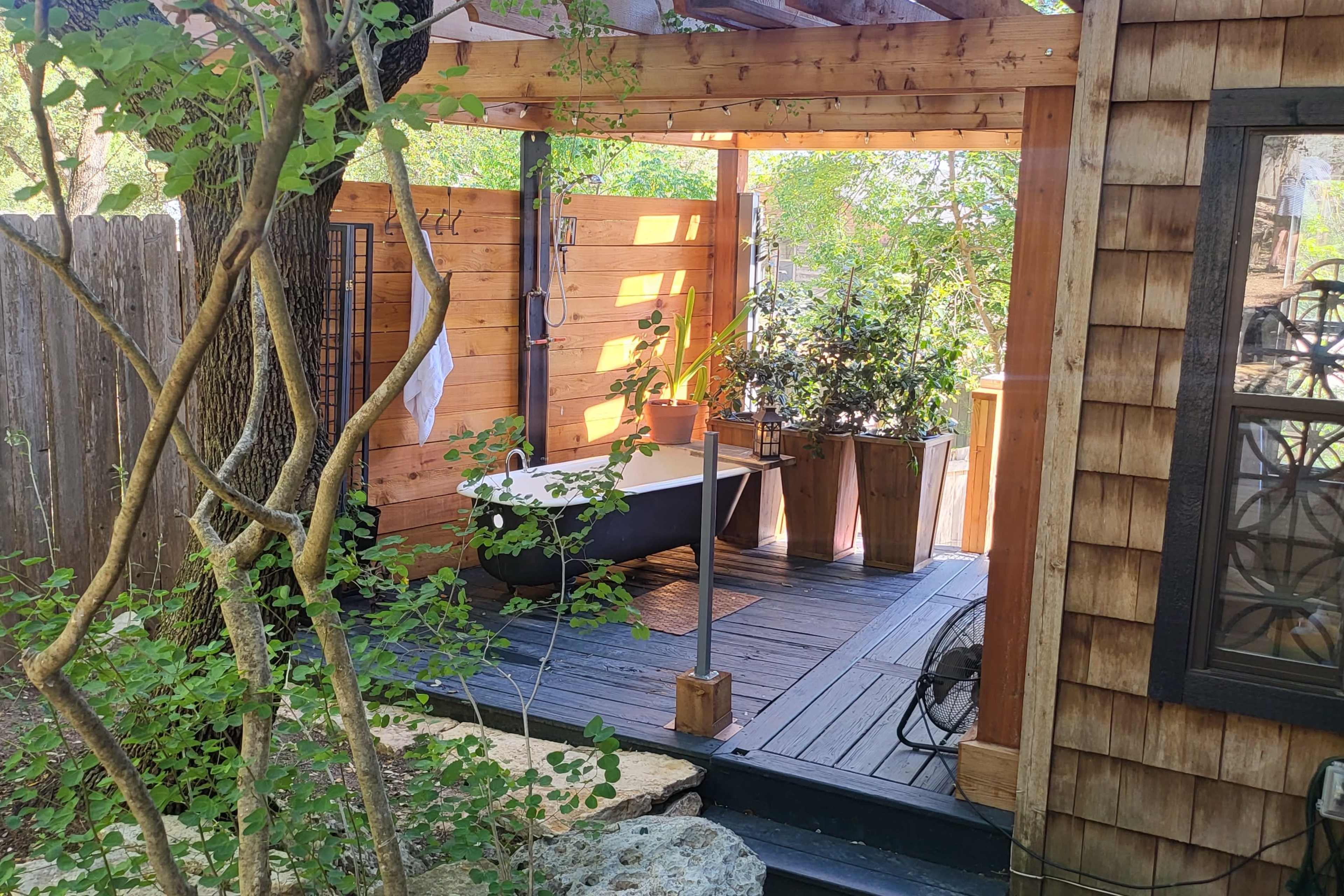 The image shows a wooden deck area featuring a freestanding bathtub surrounded by plants and trees, enclosed by wooden walls.