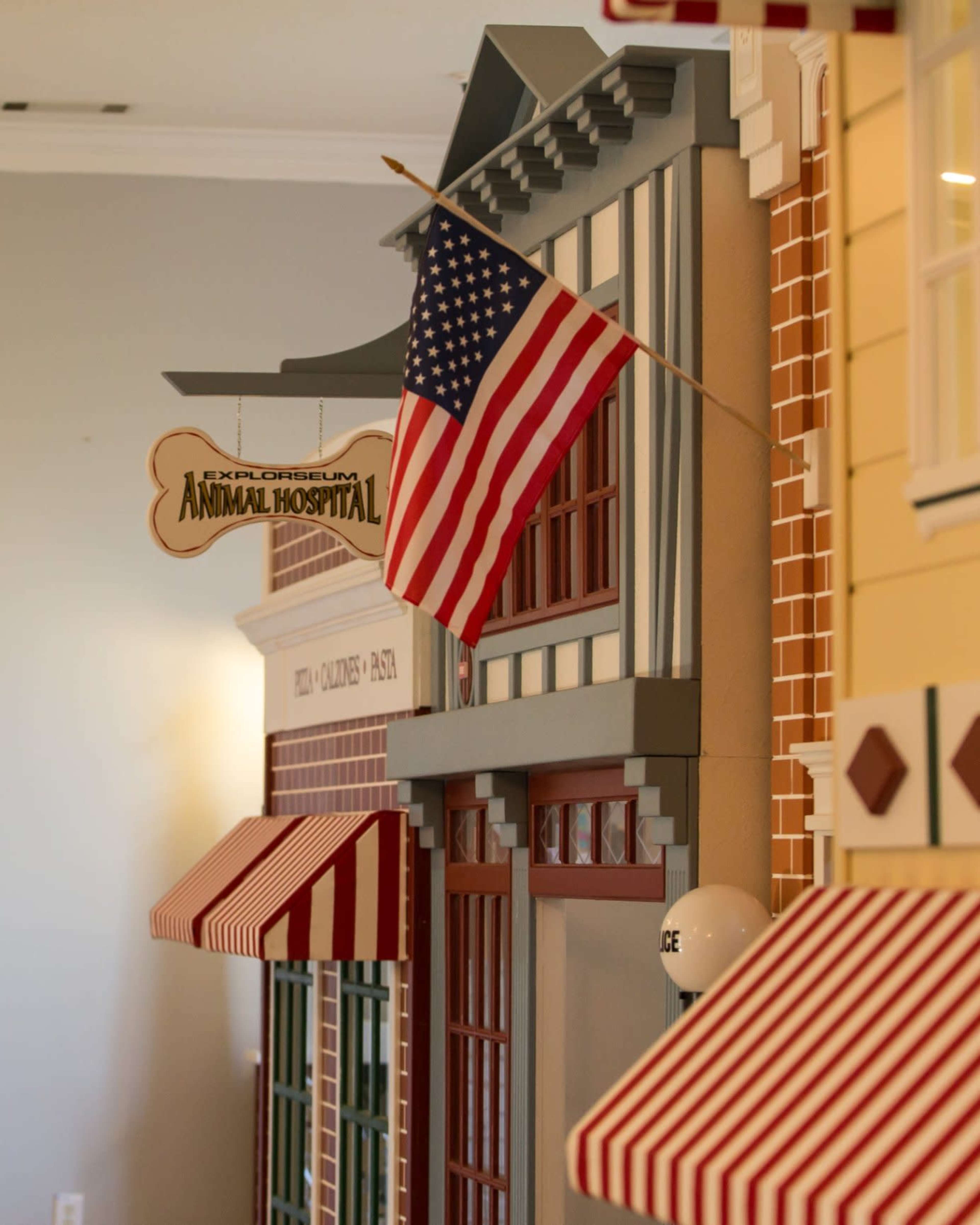 The image shows a miniature facade of a building labeled "Exploratorium Animal Hospital," featuring an American flag and striped awnings.