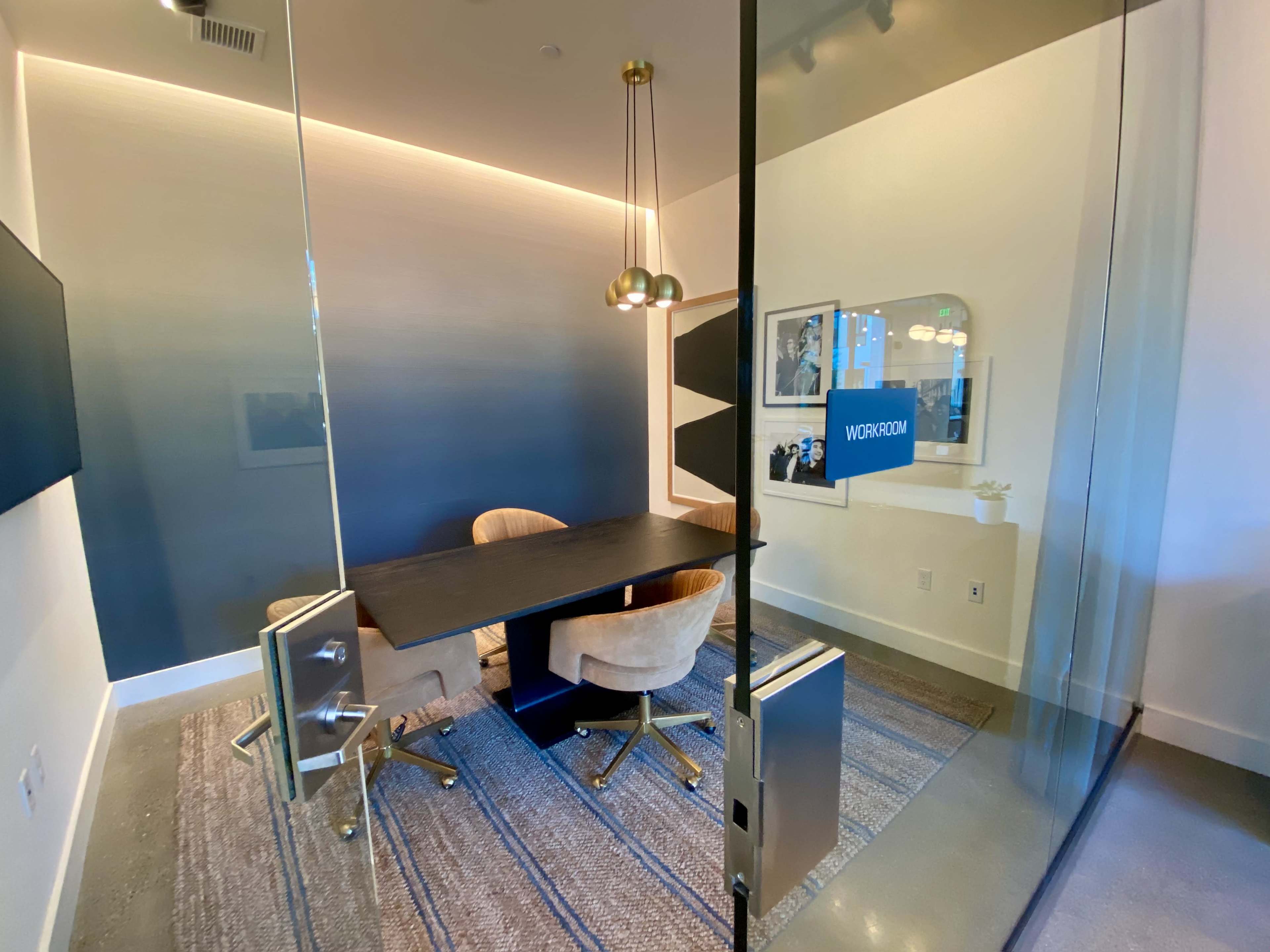A modern glass-walled meeting room with a black table and two beige chairs, illuminated by a pendant light.
