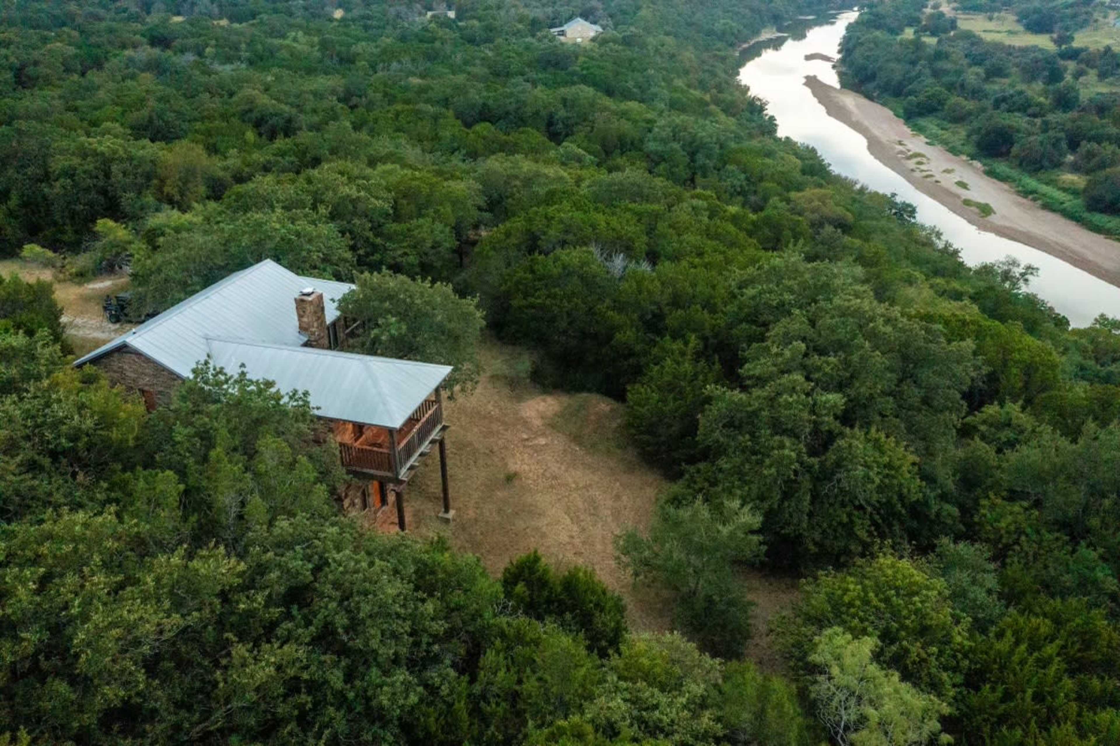 An aerial view shows a house on a hillside beside a river, surrounded by dense greenery.