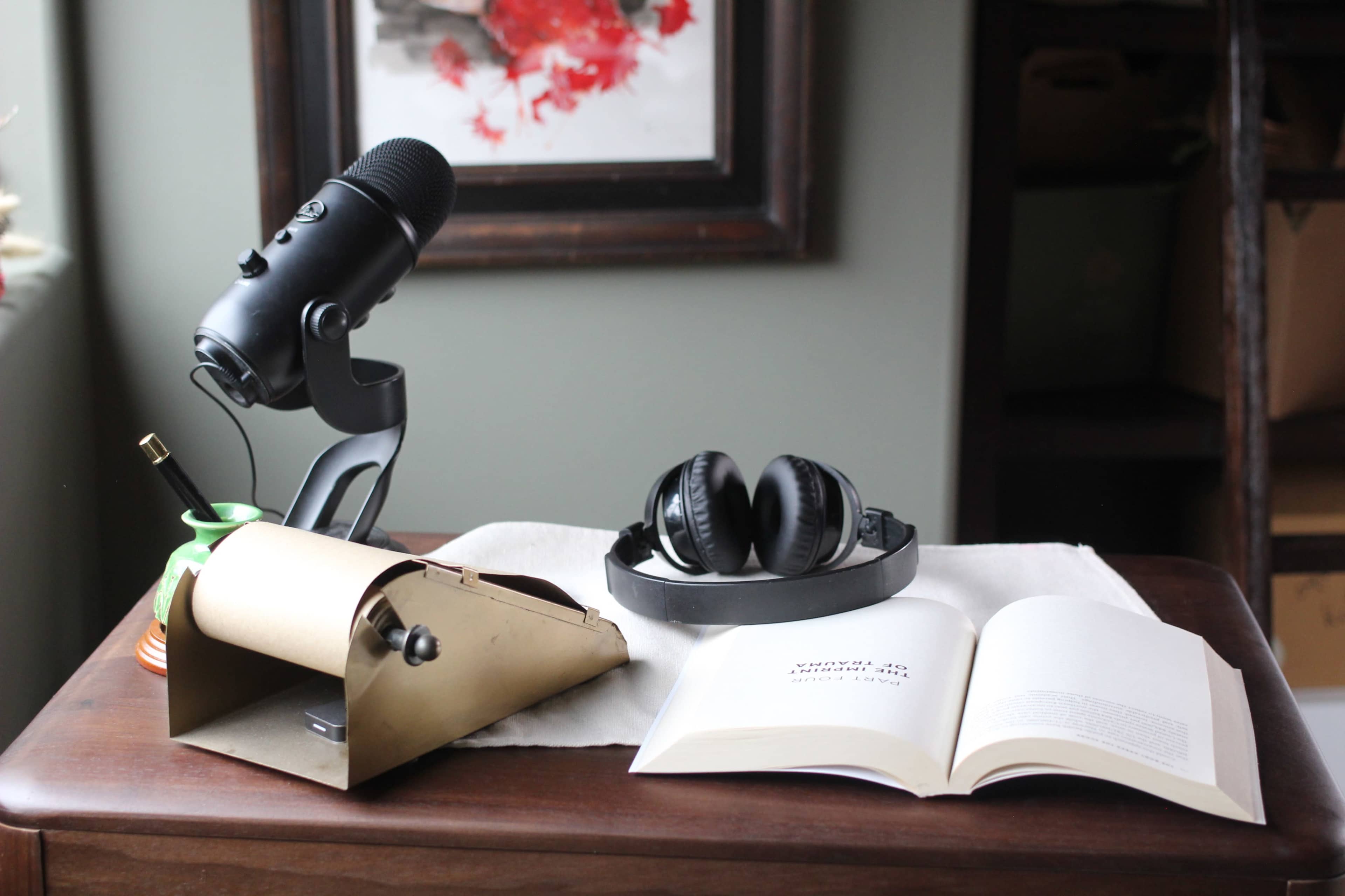 A black microphone and headphones sit on a wooden table next to an open book and a brass paper holder.