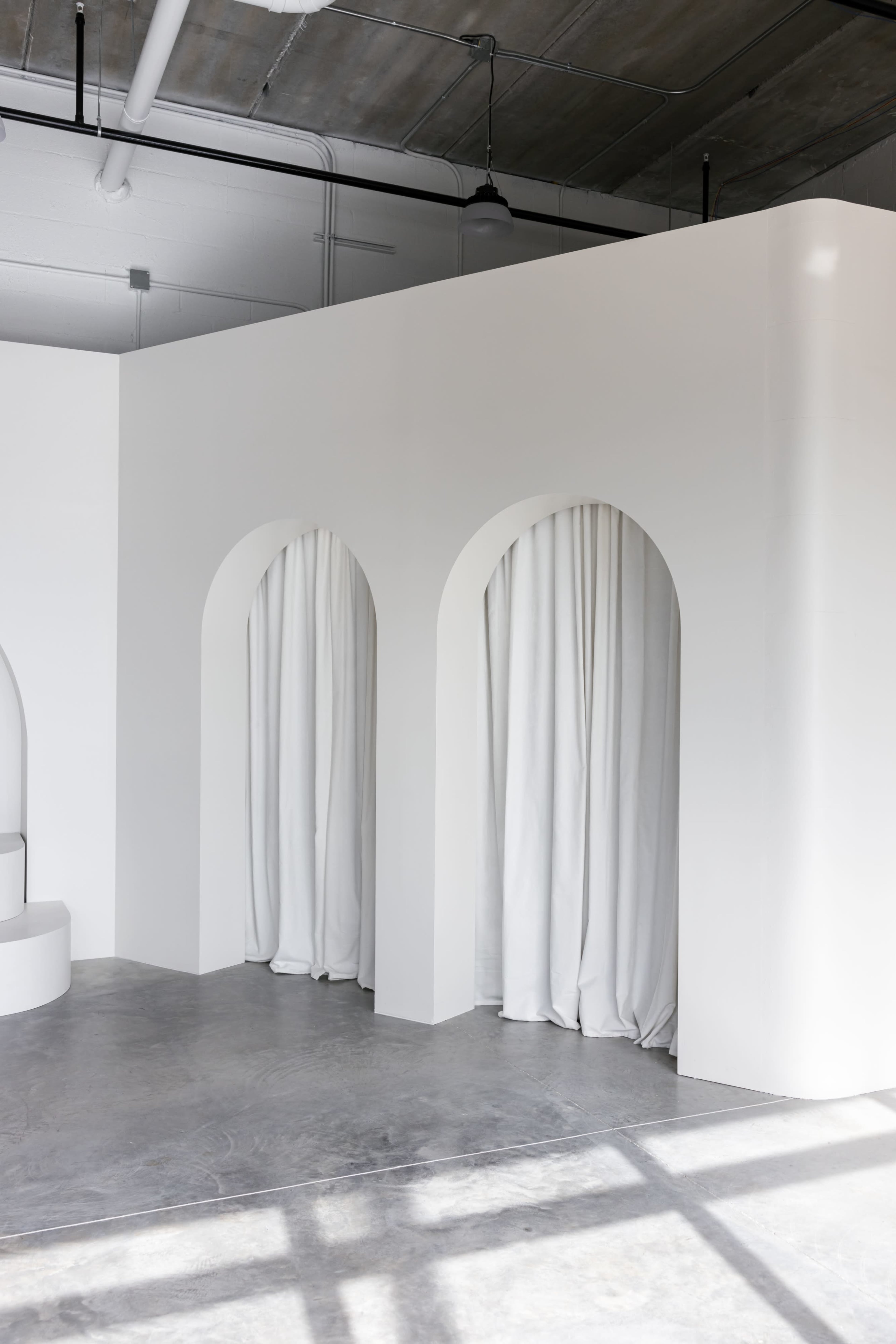 The image shows a minimalist interior space featuring three curved archways with white curtains, set against a bright white wall and a concrete floor.