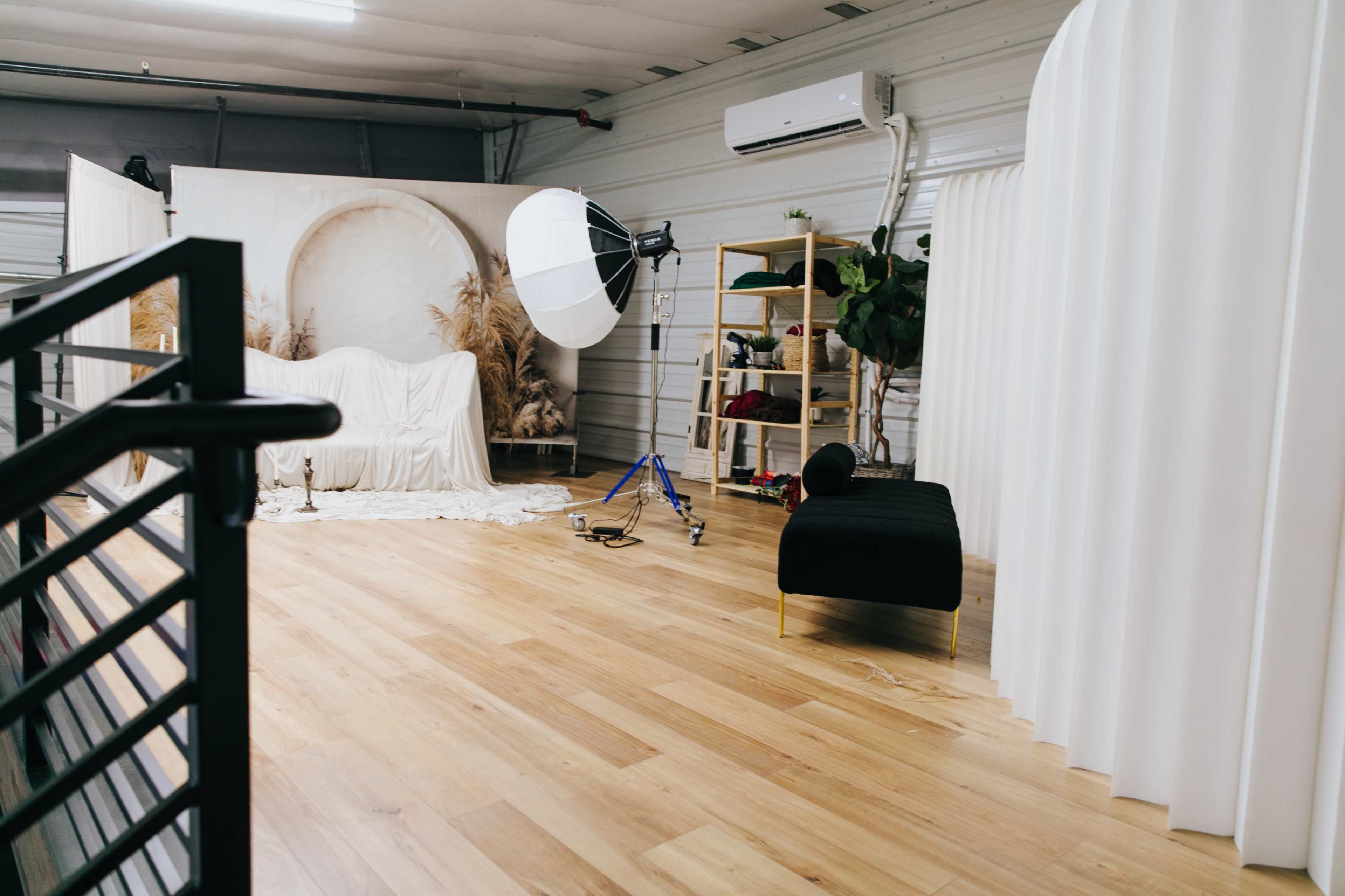 The image shows a photography studio with a light setup, a white backdrop featuring plants, a black chaise lounge, and wooden flooring.