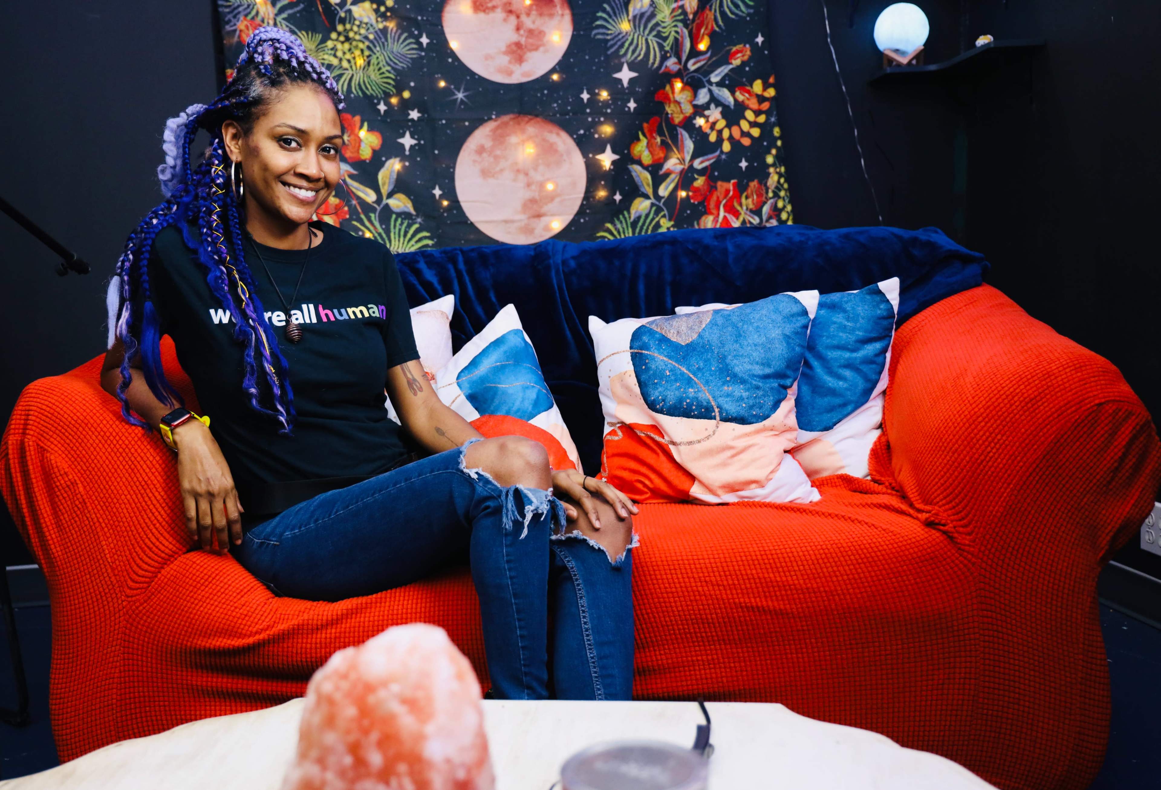 A woman with purple hair is sitting on a red sofa in a room decorated with vibrant patterns and cushions.