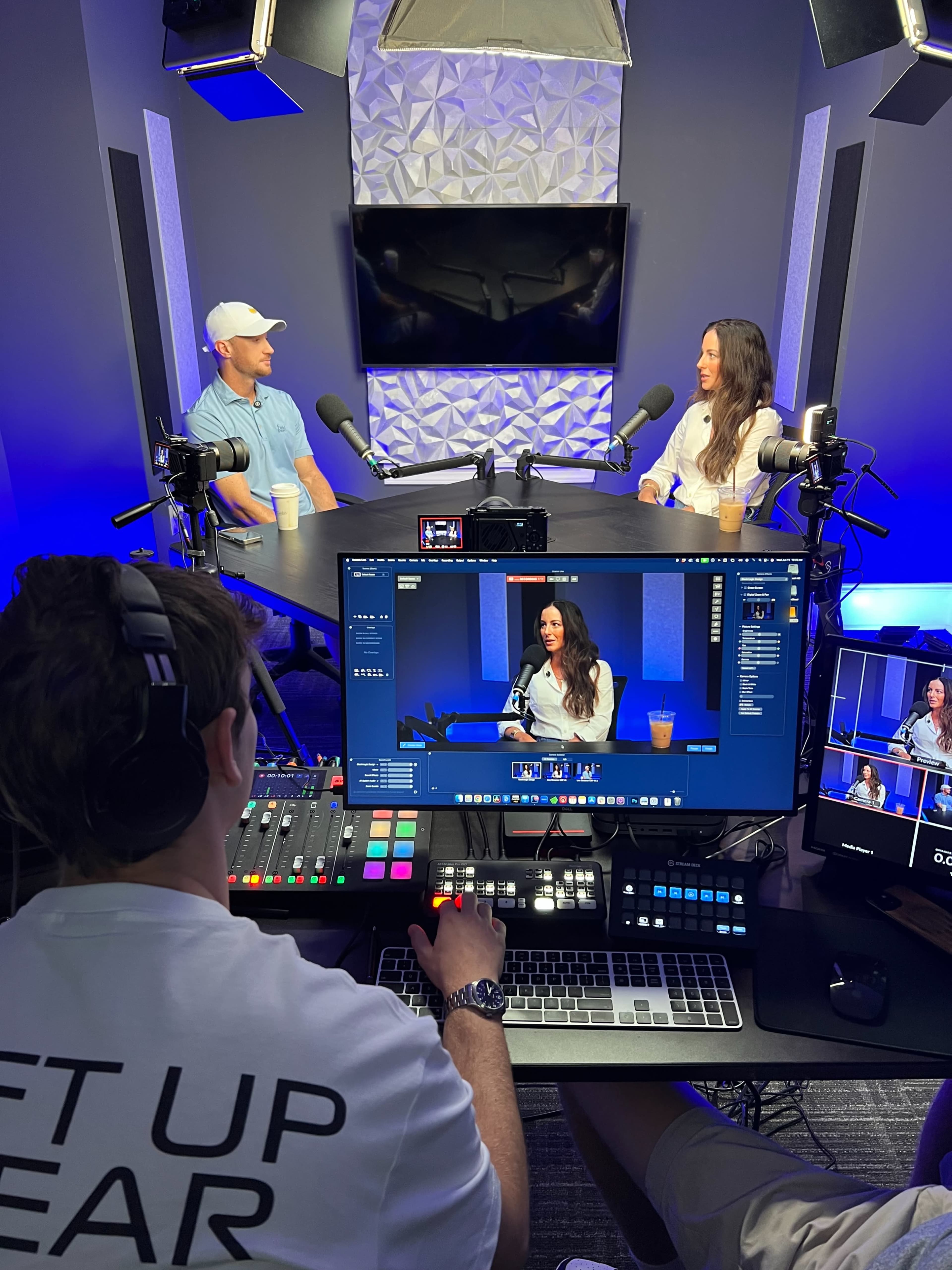 A man and a woman are seated at a table during a podcast recording, while a third person operates a camera and controls the audio equipment in a well-lit studio.