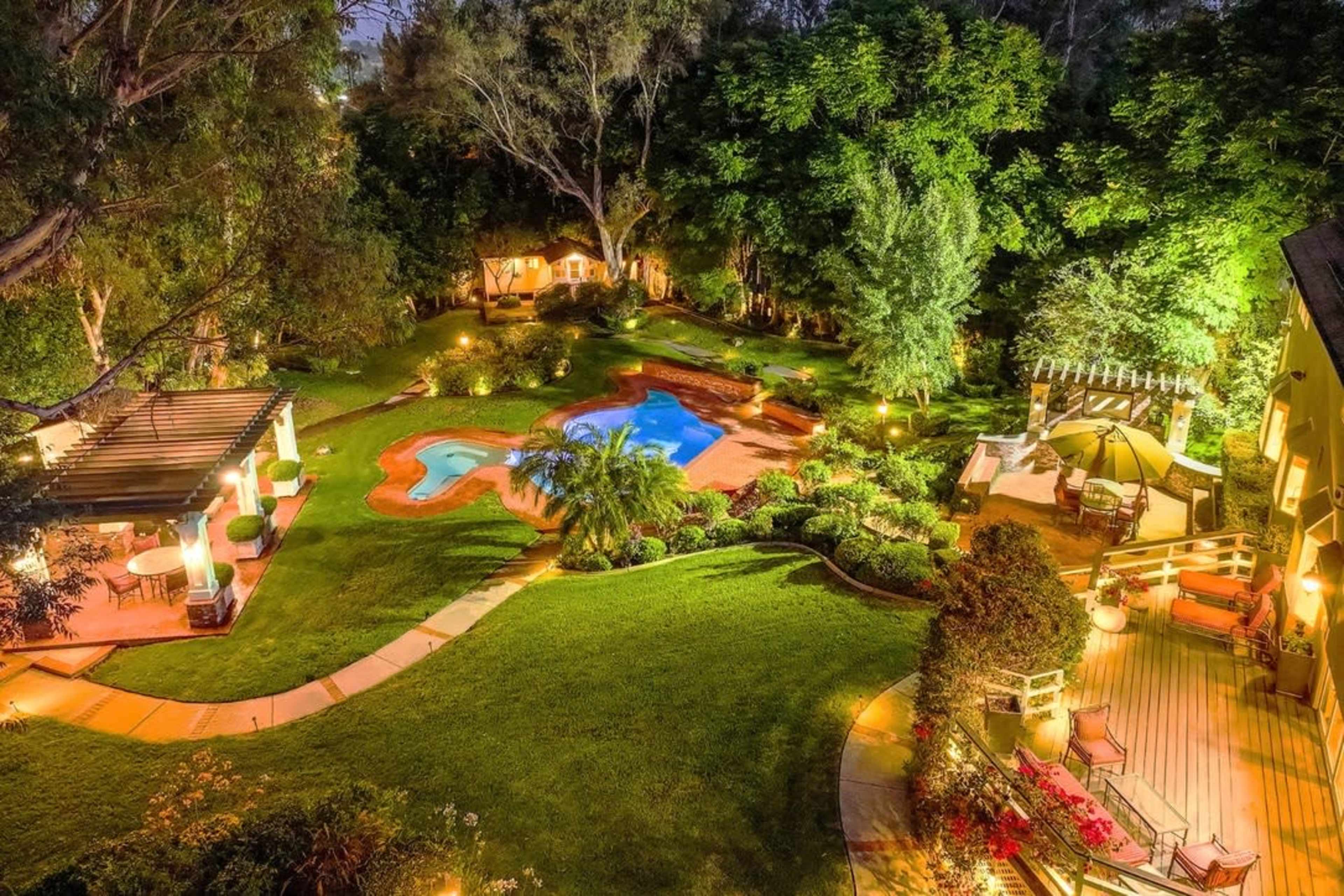 The image shows a landscaped backyard featuring a swimming pool, pathways, and seating areas surrounded by trees and gardens, illuminated at night.