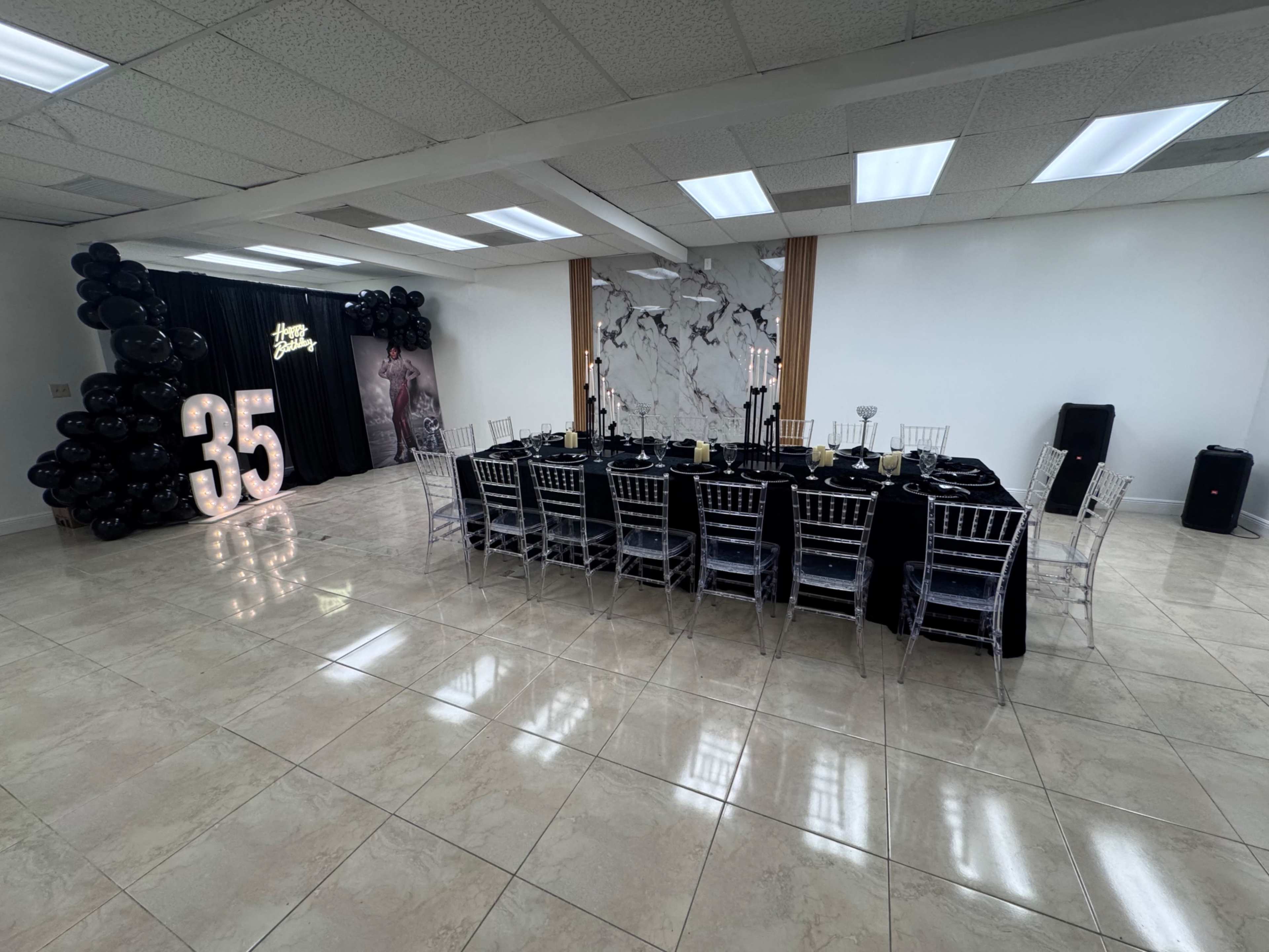 The image shows a spacious event room set up for a celebration, featuring a long banquet table with black and gold decor and a large "35" display made of lights.