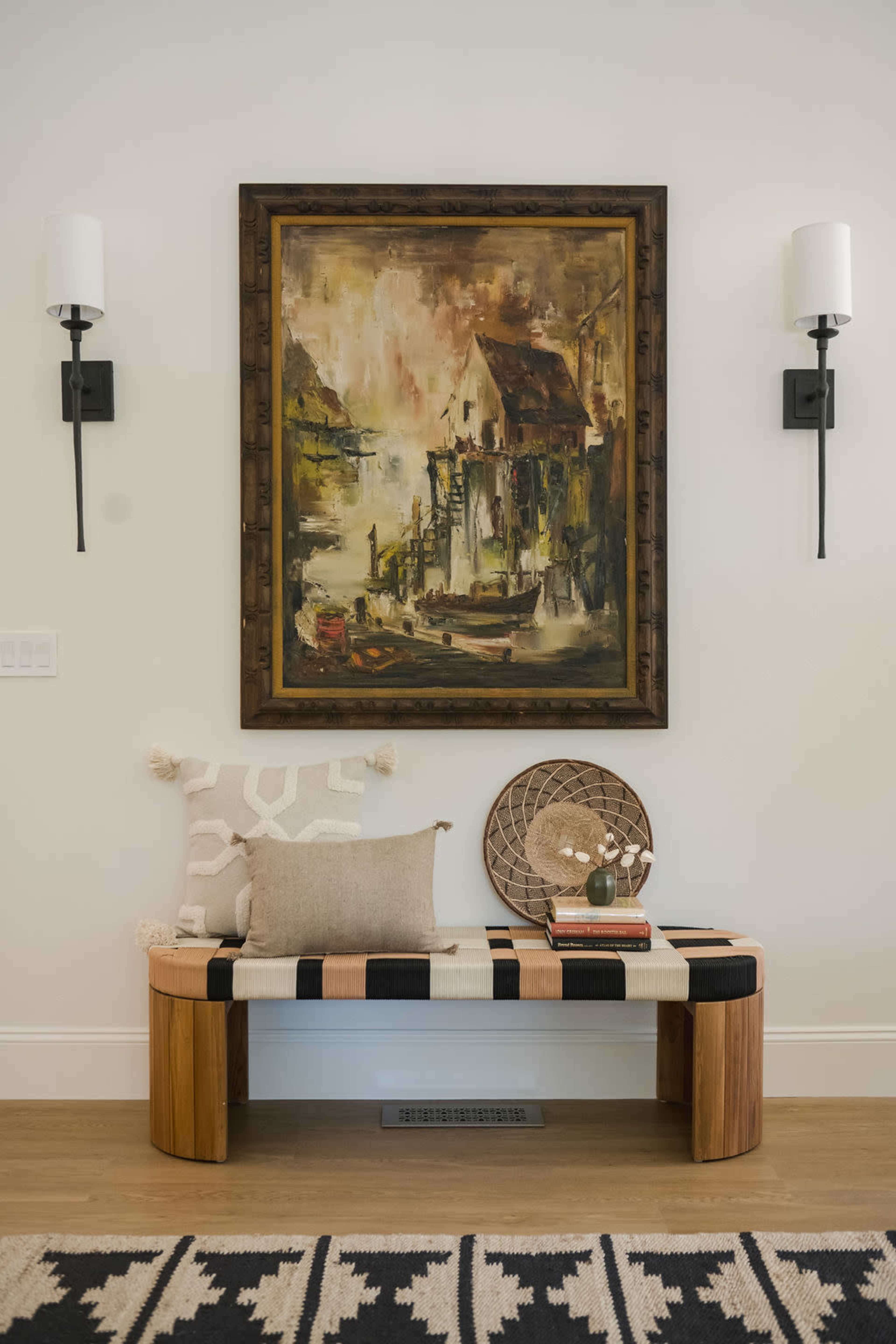 A wooden bench with patterned fabric sits beneath a large framed painting of a waterfront scene, flanked by two wall-mounted sconces.