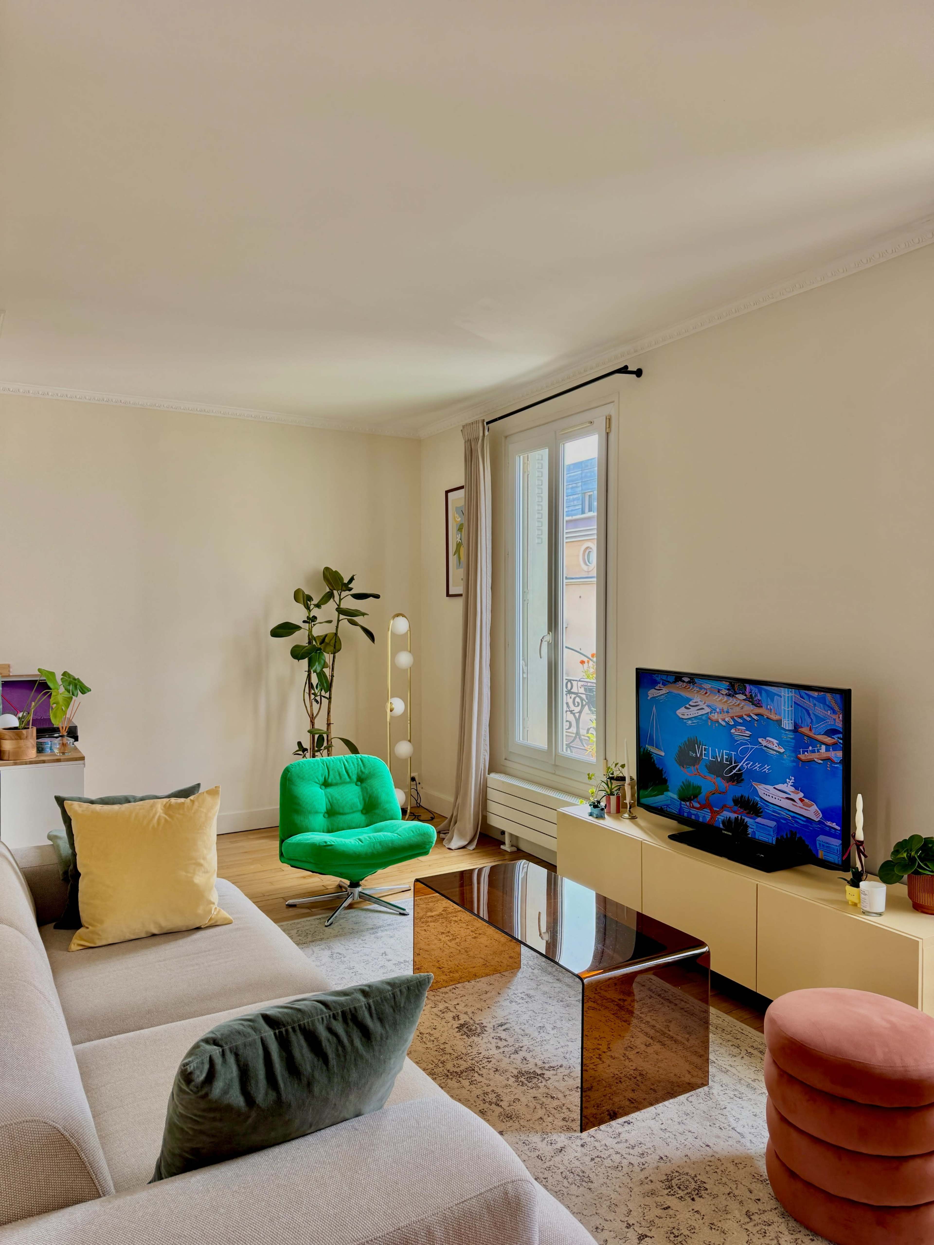 The living room features a beige sofa, a green accent chair, a glass coffee table, and a television displaying colorful content, with a window letting in natural light.