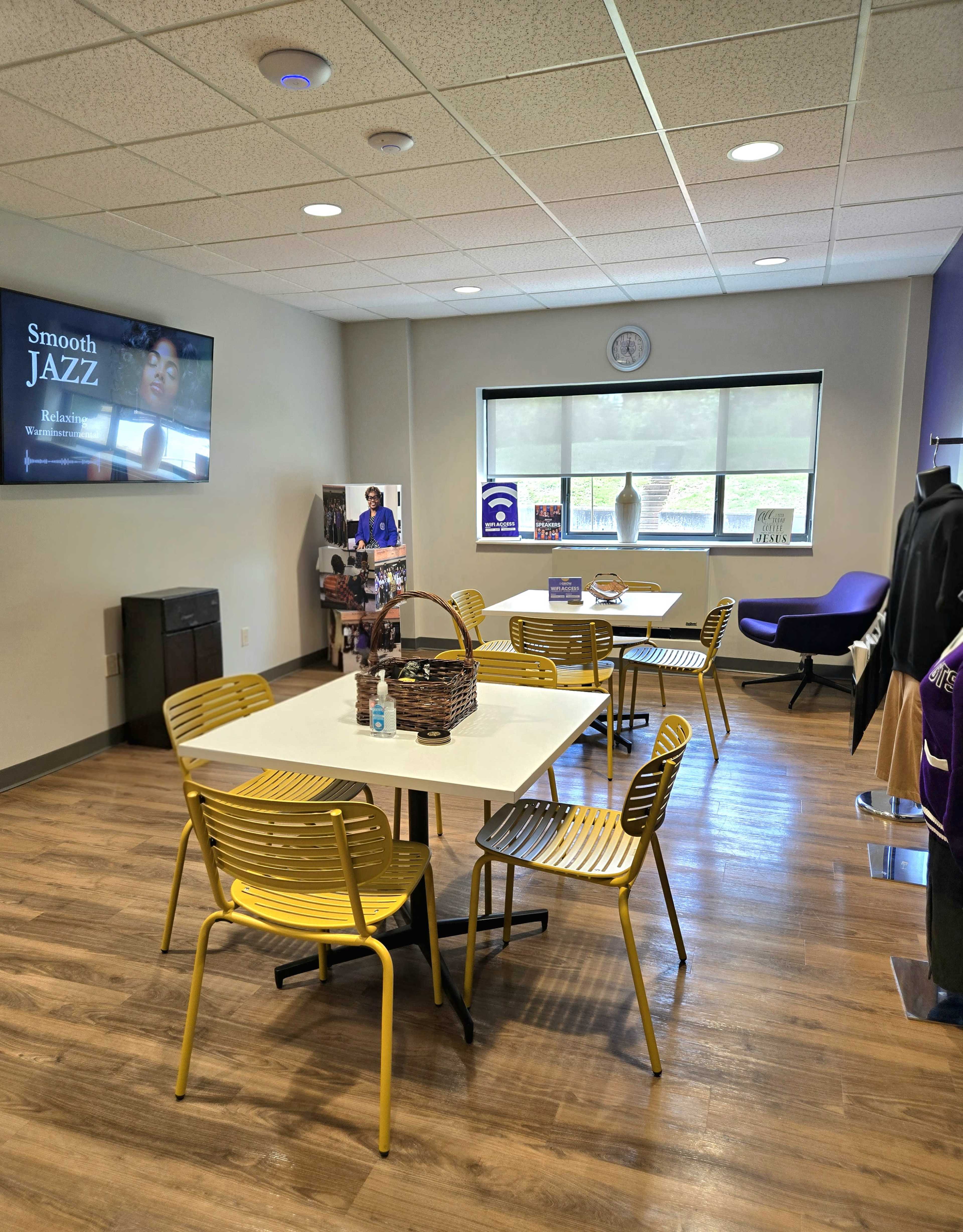 The image shows a modern room with multiple tables and chairs, featuring a television screen on the wall and various displays, including a coat and informational materials.
