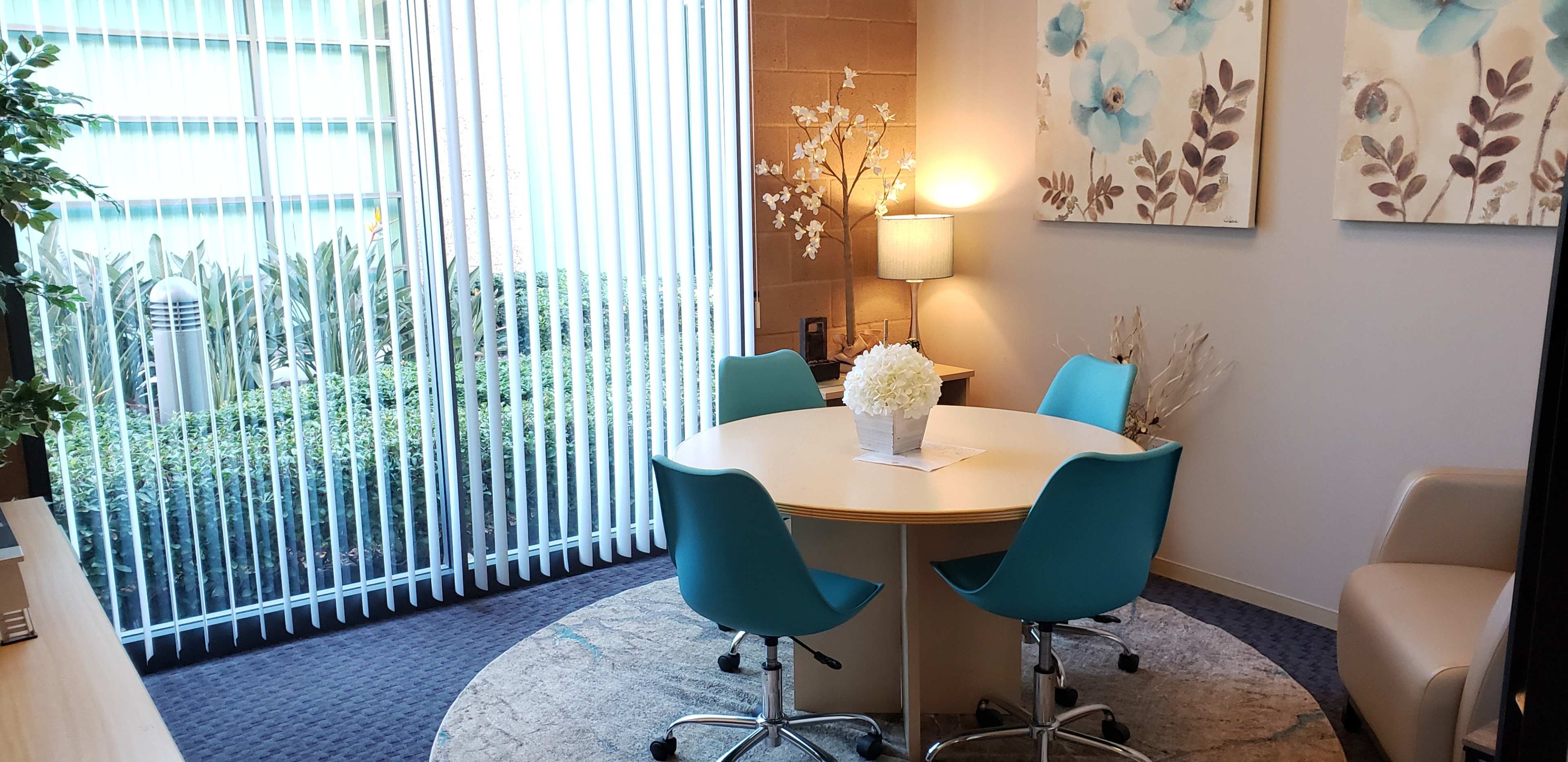 A small meeting room with a round table and four blue chairs, featuring a window with vertical blinds and a view of green plants outside.