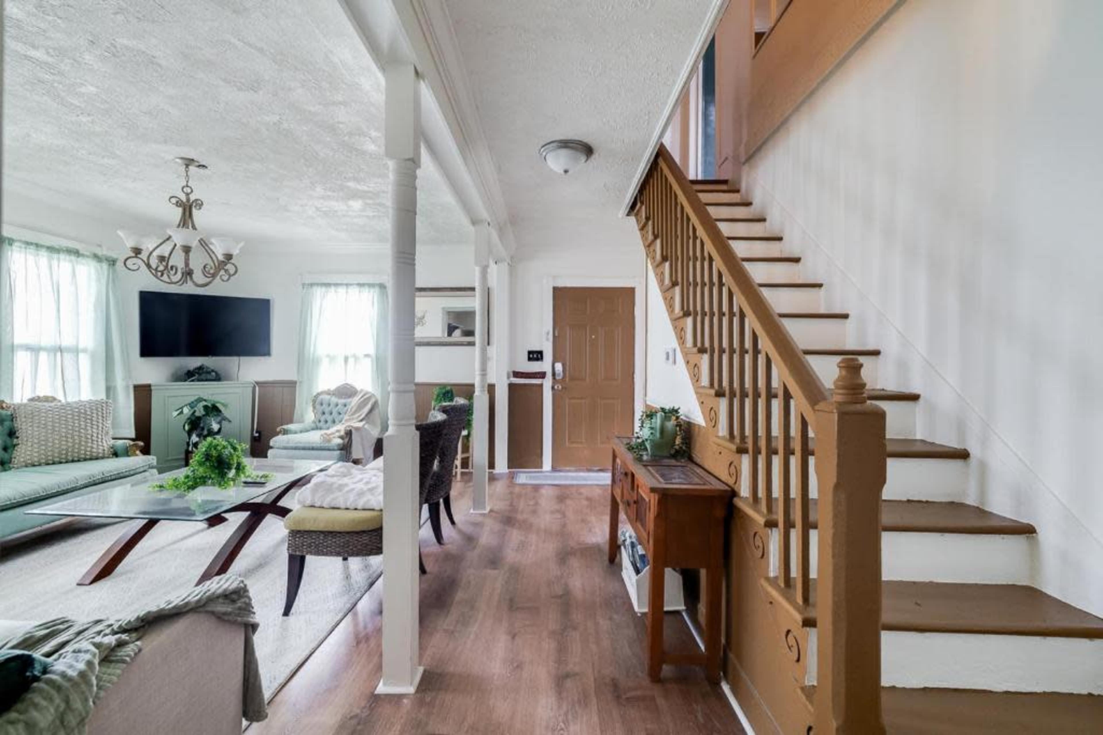 A well-lit living area with a staircase, a coffee table, and various seating arrangements, leading to a front door.