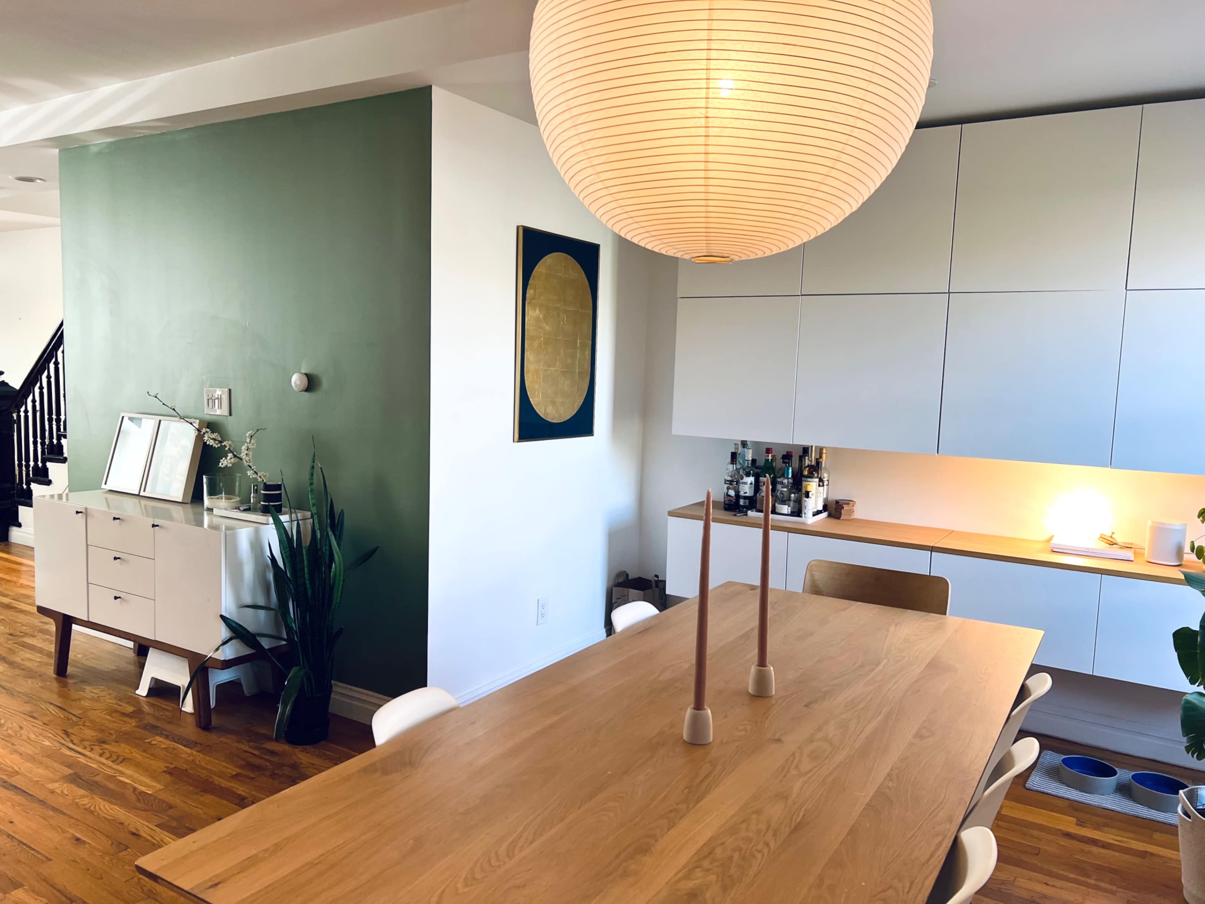 A modern dining area features a large wooden table surrounded by white chairs, with a pendant light overhead and a green accent wall in the background.