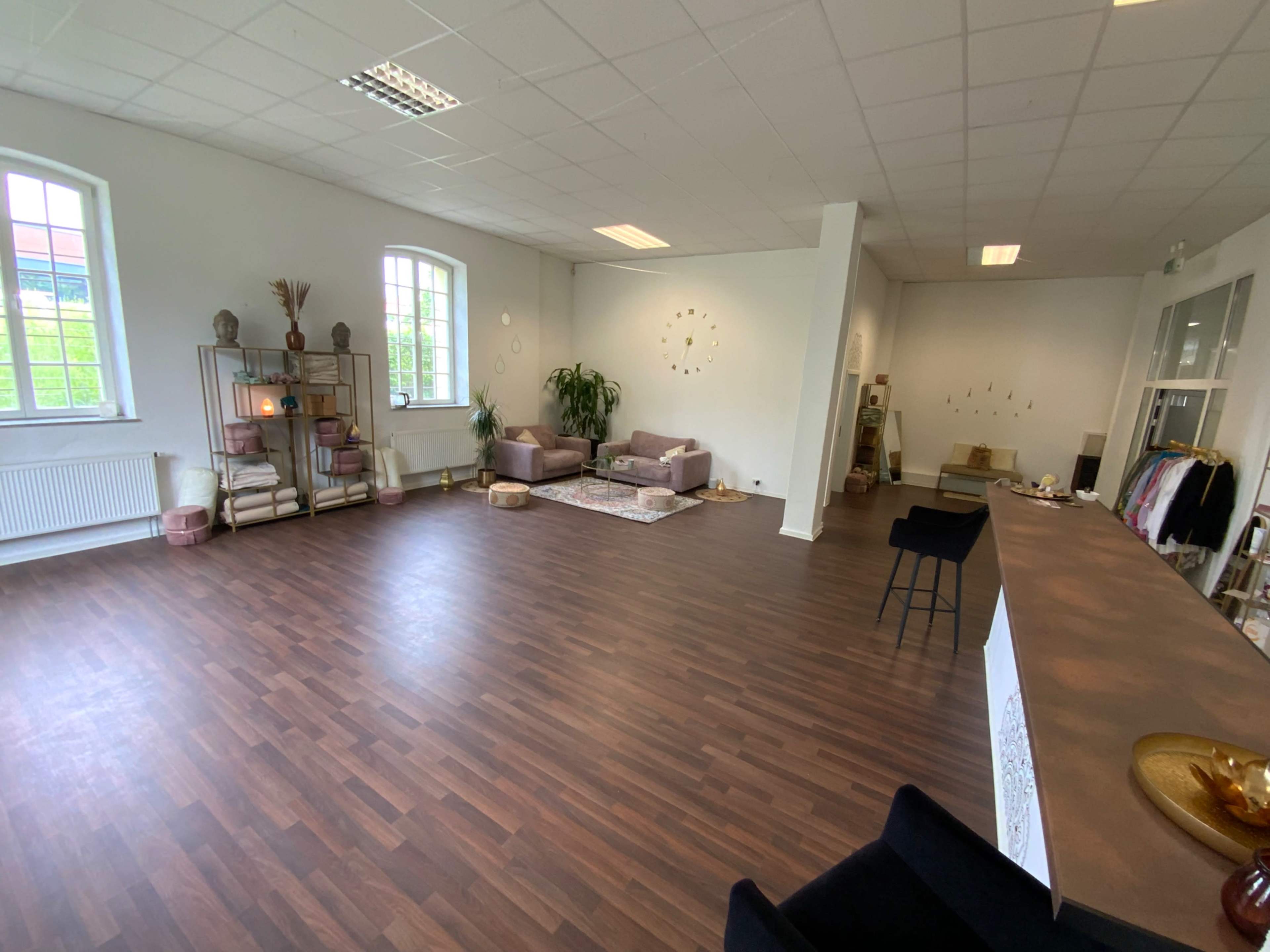 The image shows a spacious interior room with hardwood flooring, featuring two sofas, a coffee table, plants, and a long counter with chairs along one side.