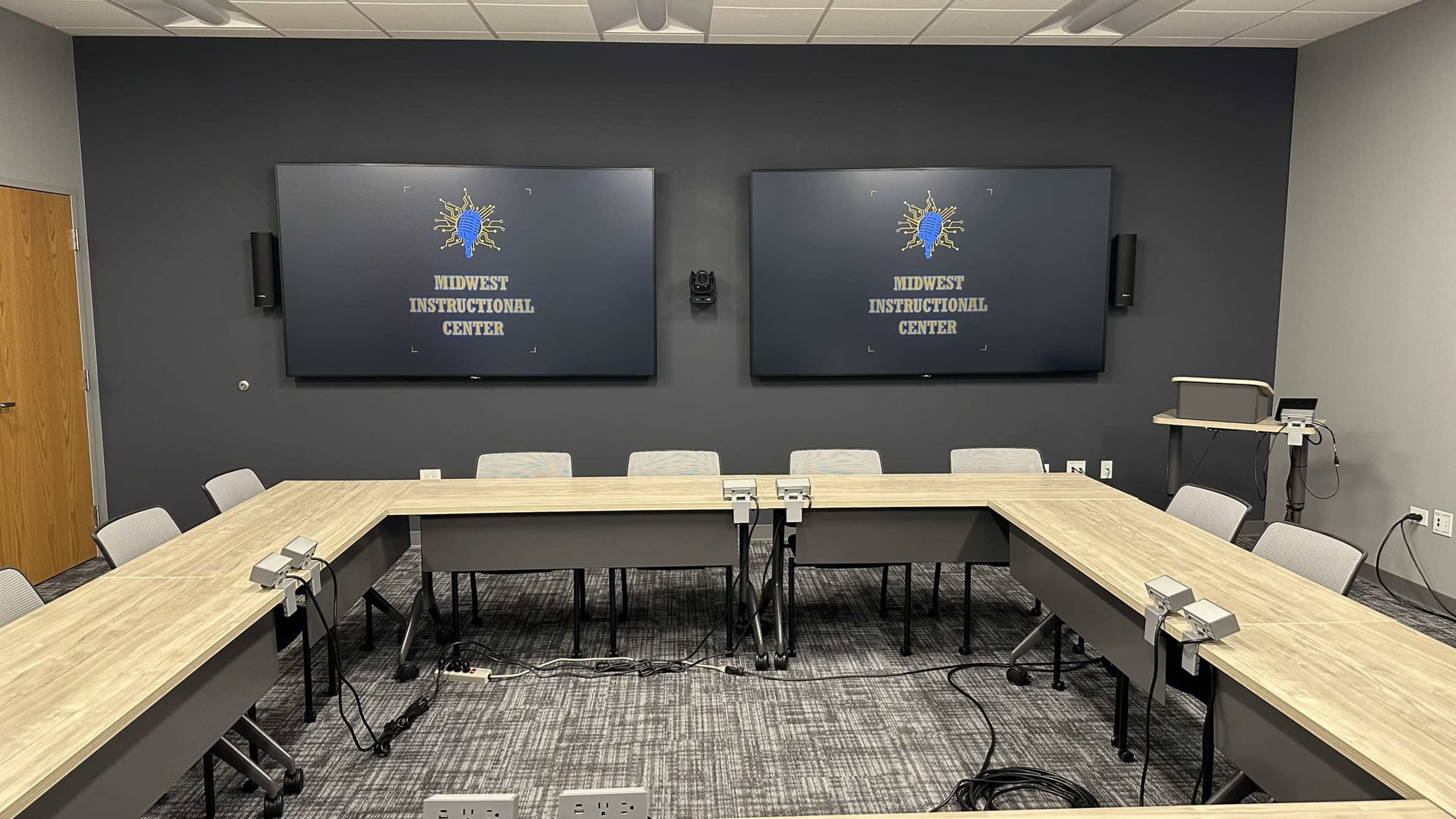 The image shows a modern conference room equipped with two large screens and a rectangular table surrounded by multiple chairs.