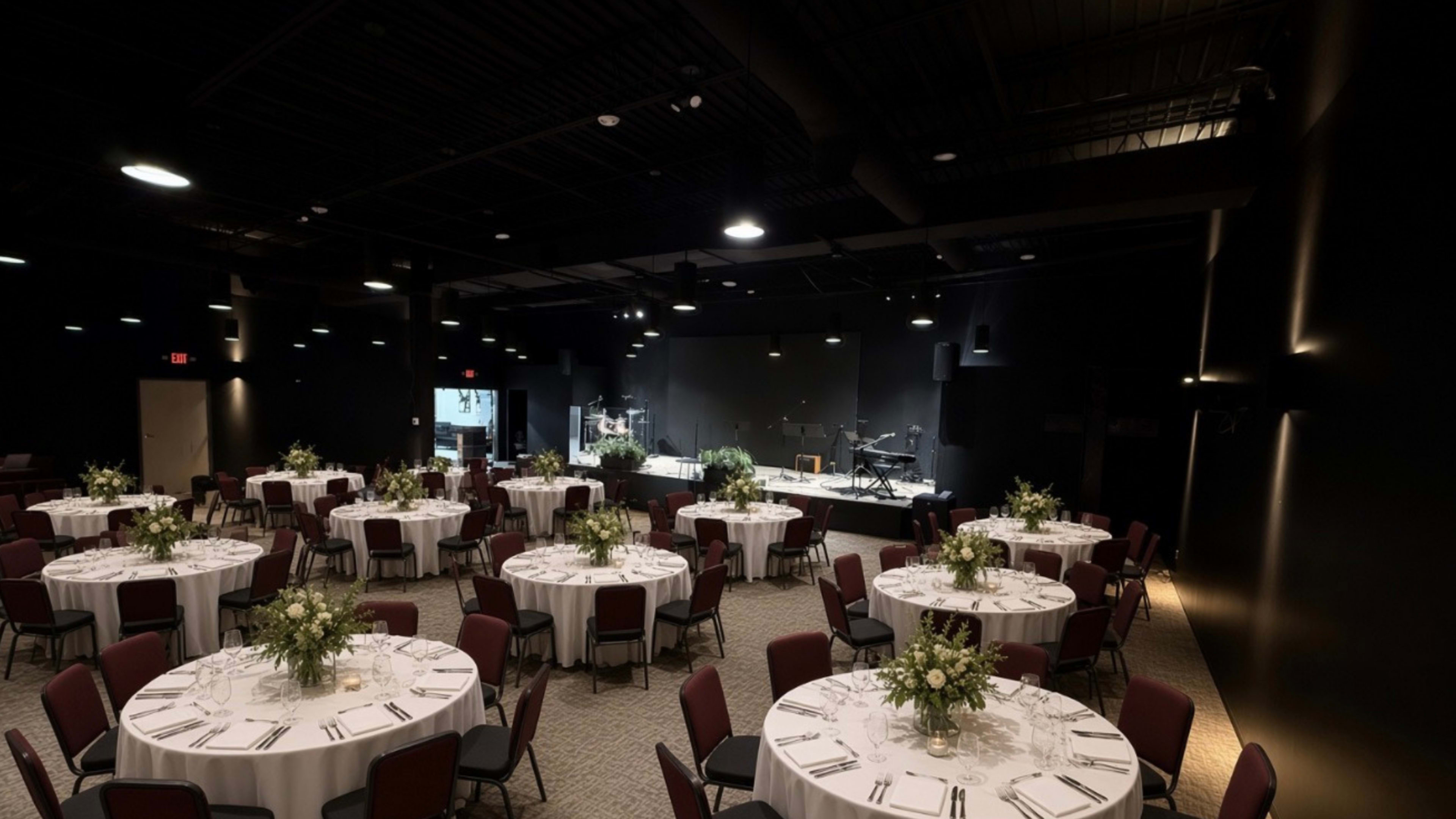 The image shows a dimly lit event space with several round tables set with white tablecloths and floral centerpieces, facing a small stage equipped with musical instruments.