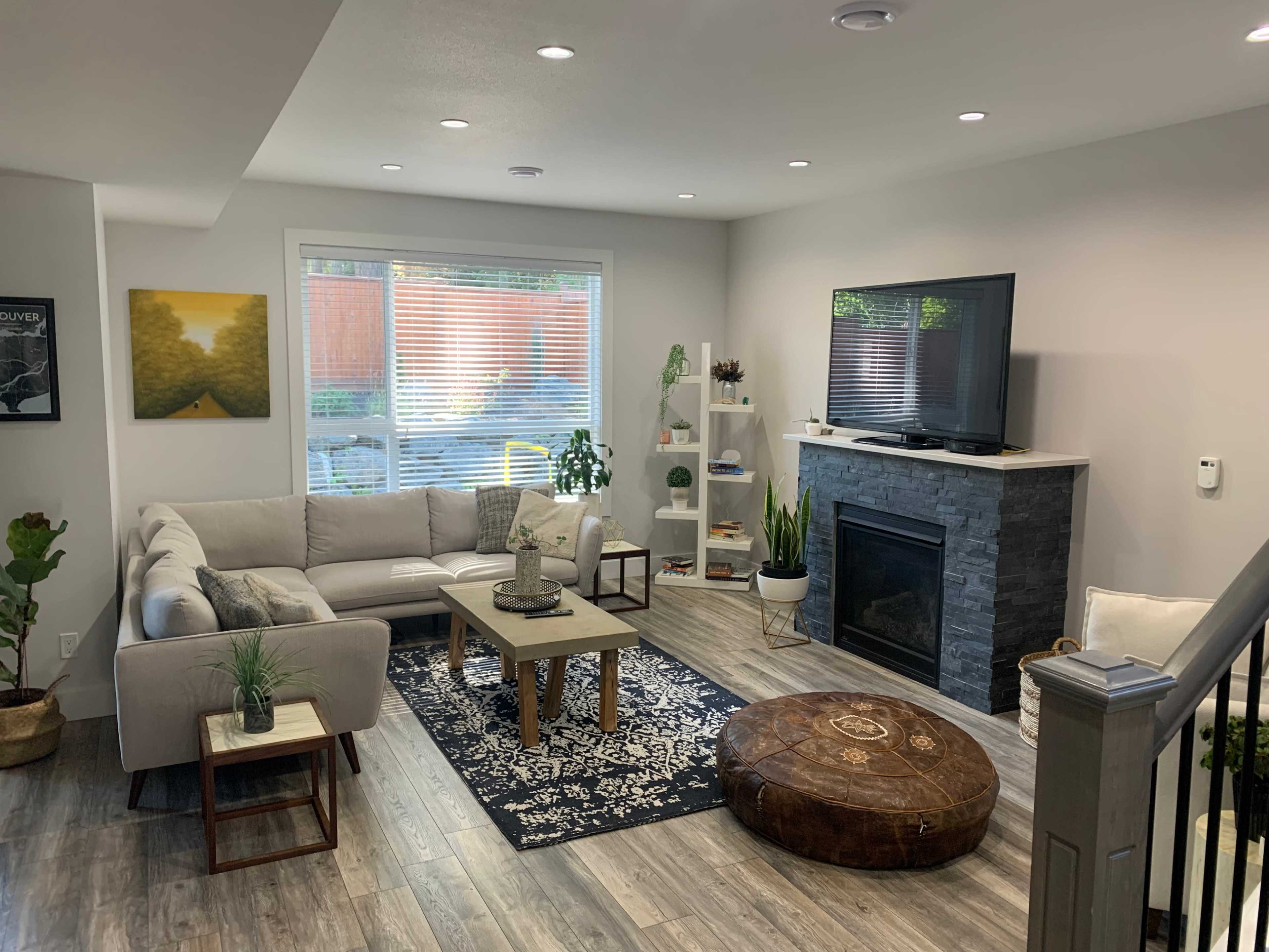 A modern living room with a sectional sofa, a coffee table, a fireplace, a flat-screen TV, and decorative plants.