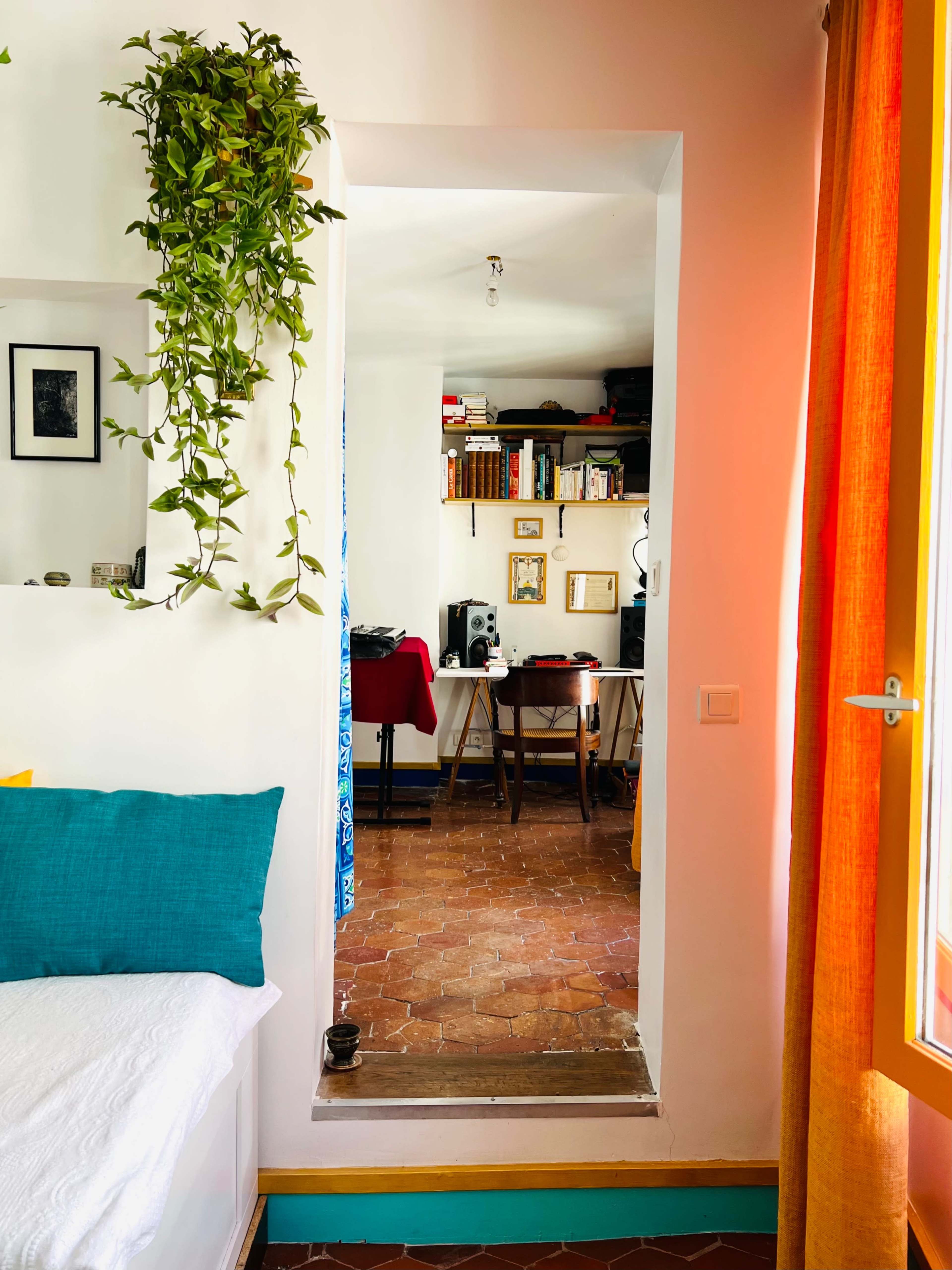 A potted plant hangs beside an archway leading into a room with a desk, bookshelves, and a red table.