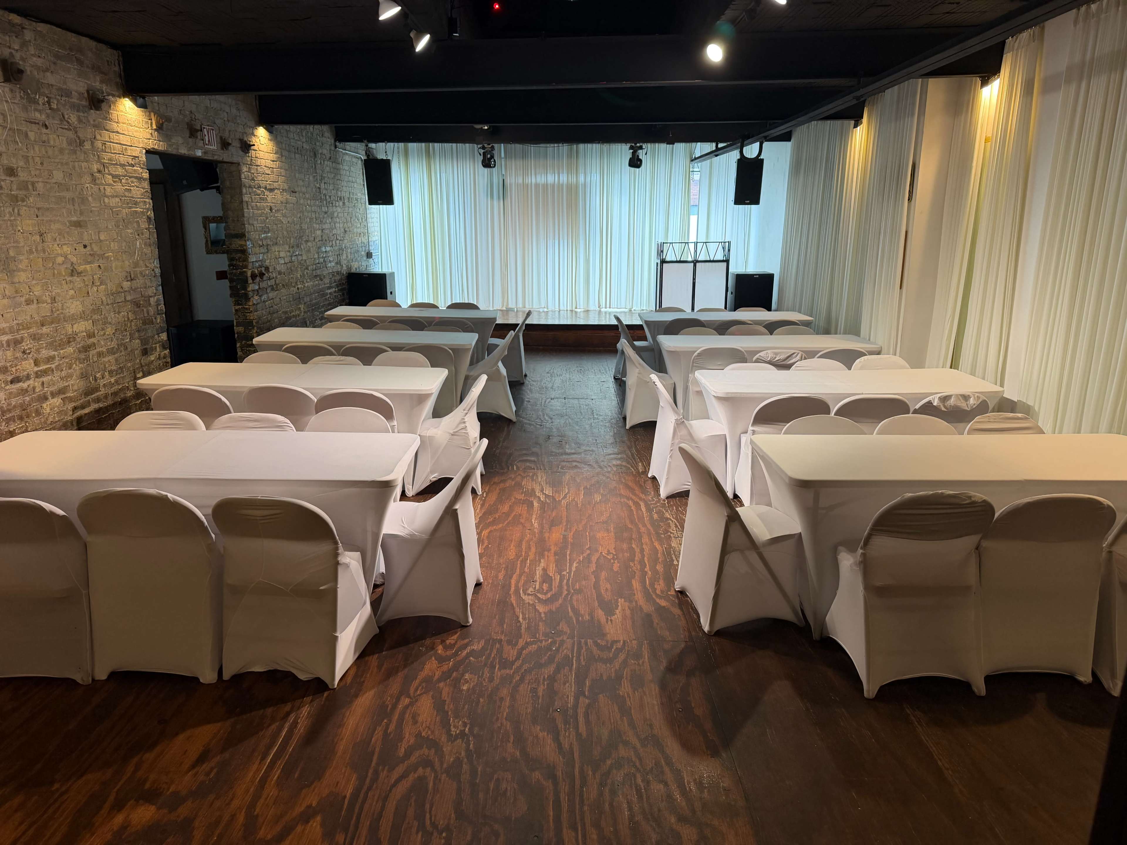 The image shows a spacious event venue set up with rows of neatly arranged tables covered in white tablecloths, surrounded by wooden flooring and soft drapery.