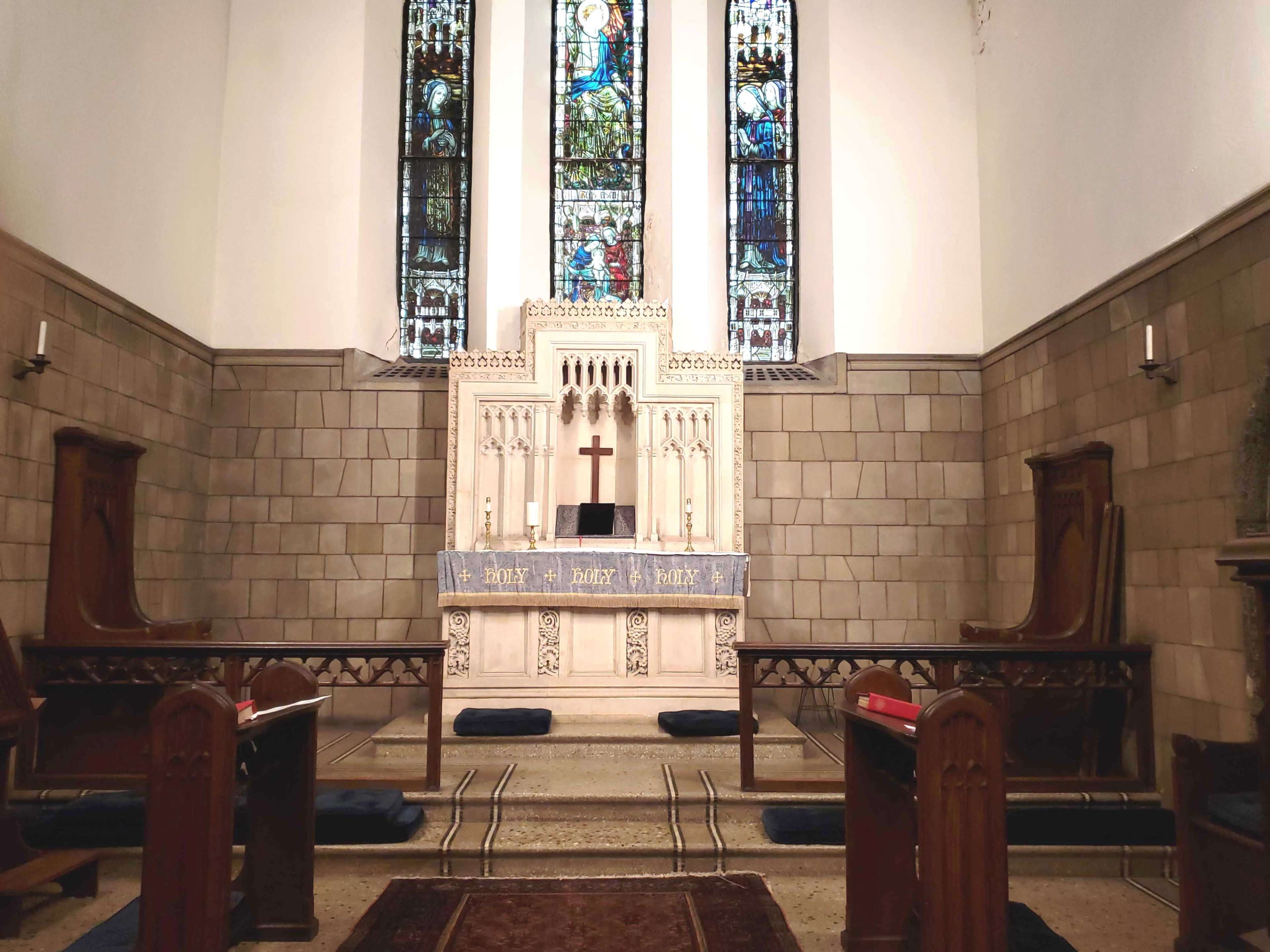 The image shows a church altar with a cross, flanked by stained glass windows, surrounded by wooden benches and a decorative rug.