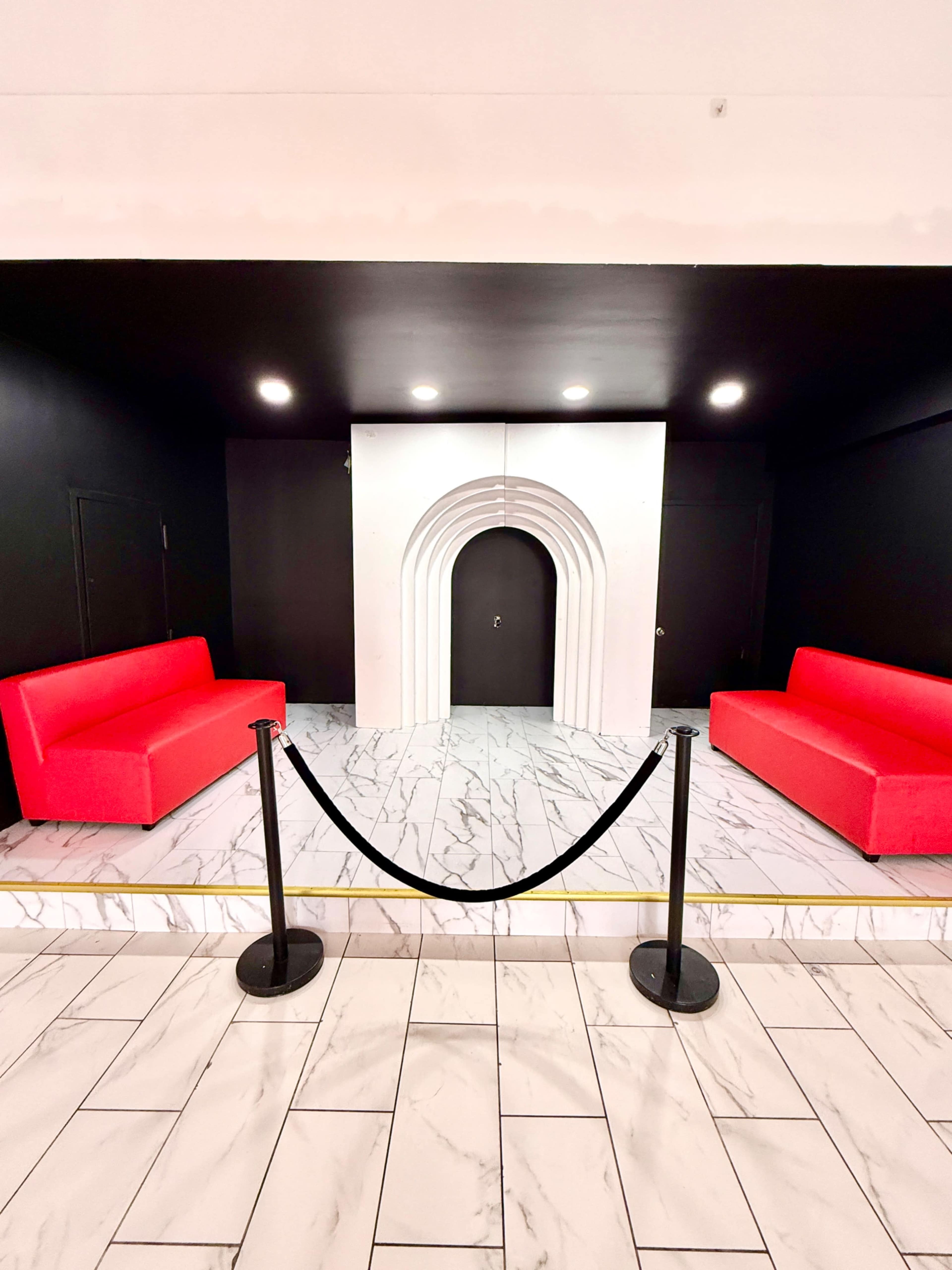 The image shows a minimalist interior with two red sofas facing a white archway leading to a dark doorway, surrounded by a black wall and marble tile flooring.