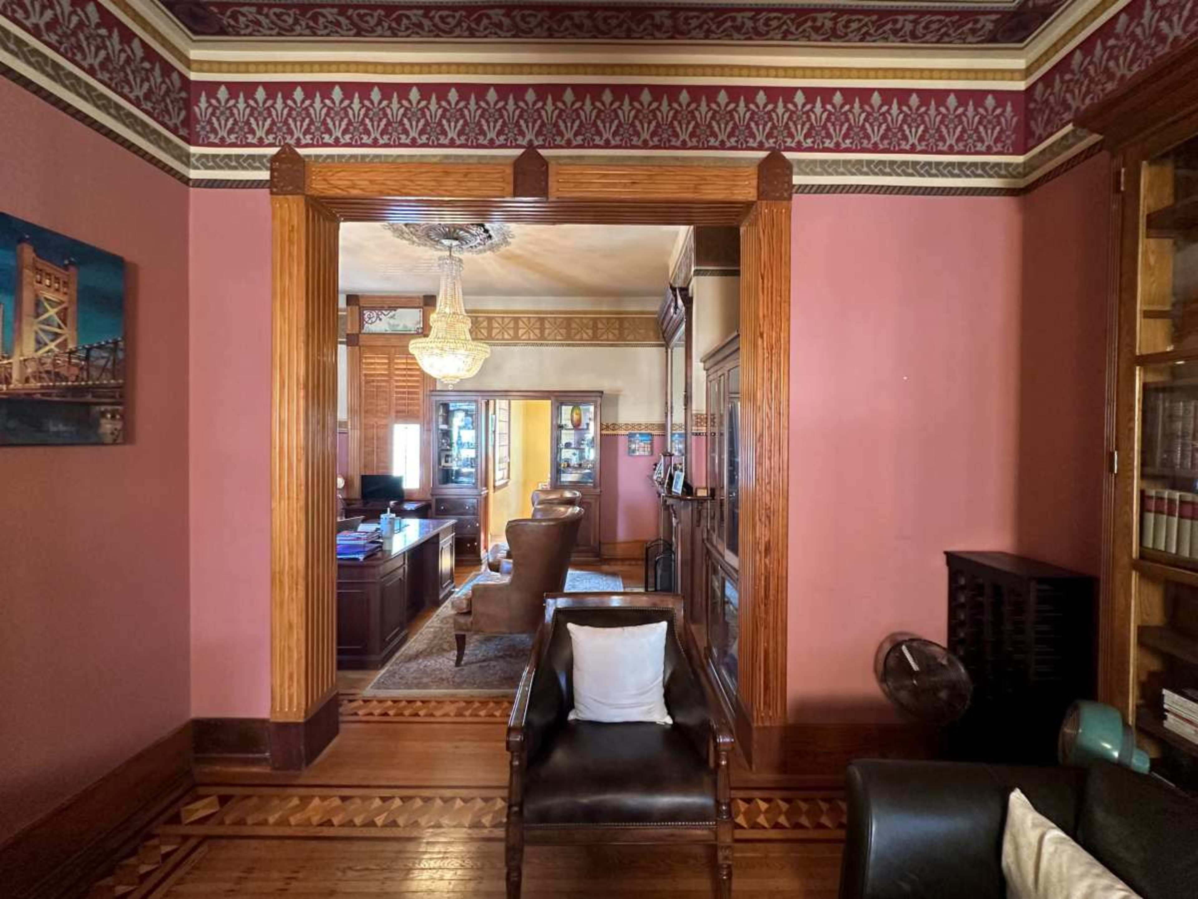 The image depicts a richly decorated room with wooden trim and ornamental wallpaper, featuring a doorway that opens to an office area with a desk and a comfortable chair.