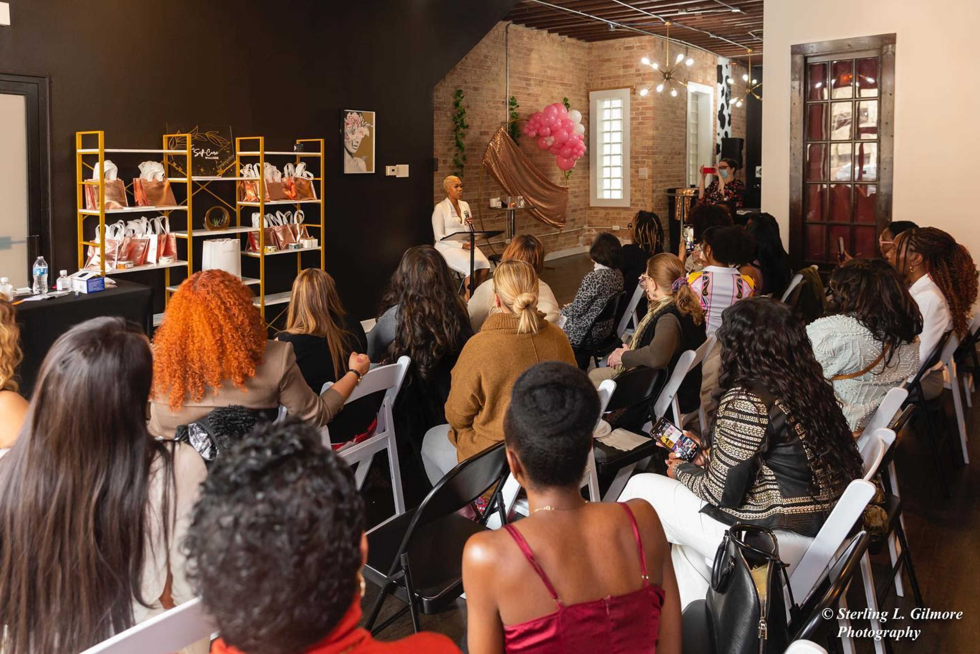 A crowd of women sits facing a speaker in a modern event space adorned with pink decorations and product displays.