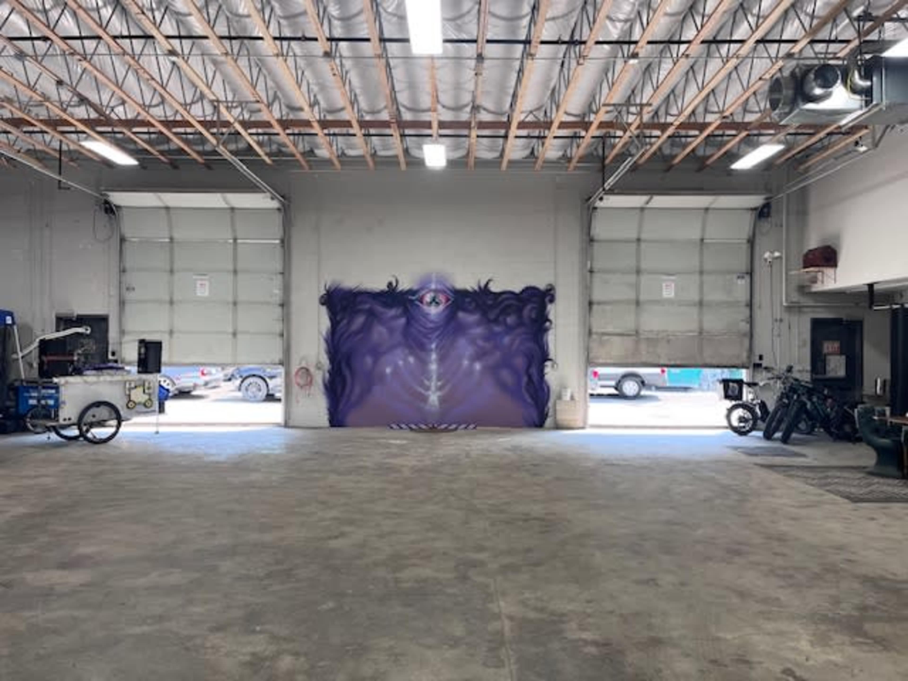 The interior of a large, empty warehouse with a prominent purple mural featuring an eye on one of the walls.