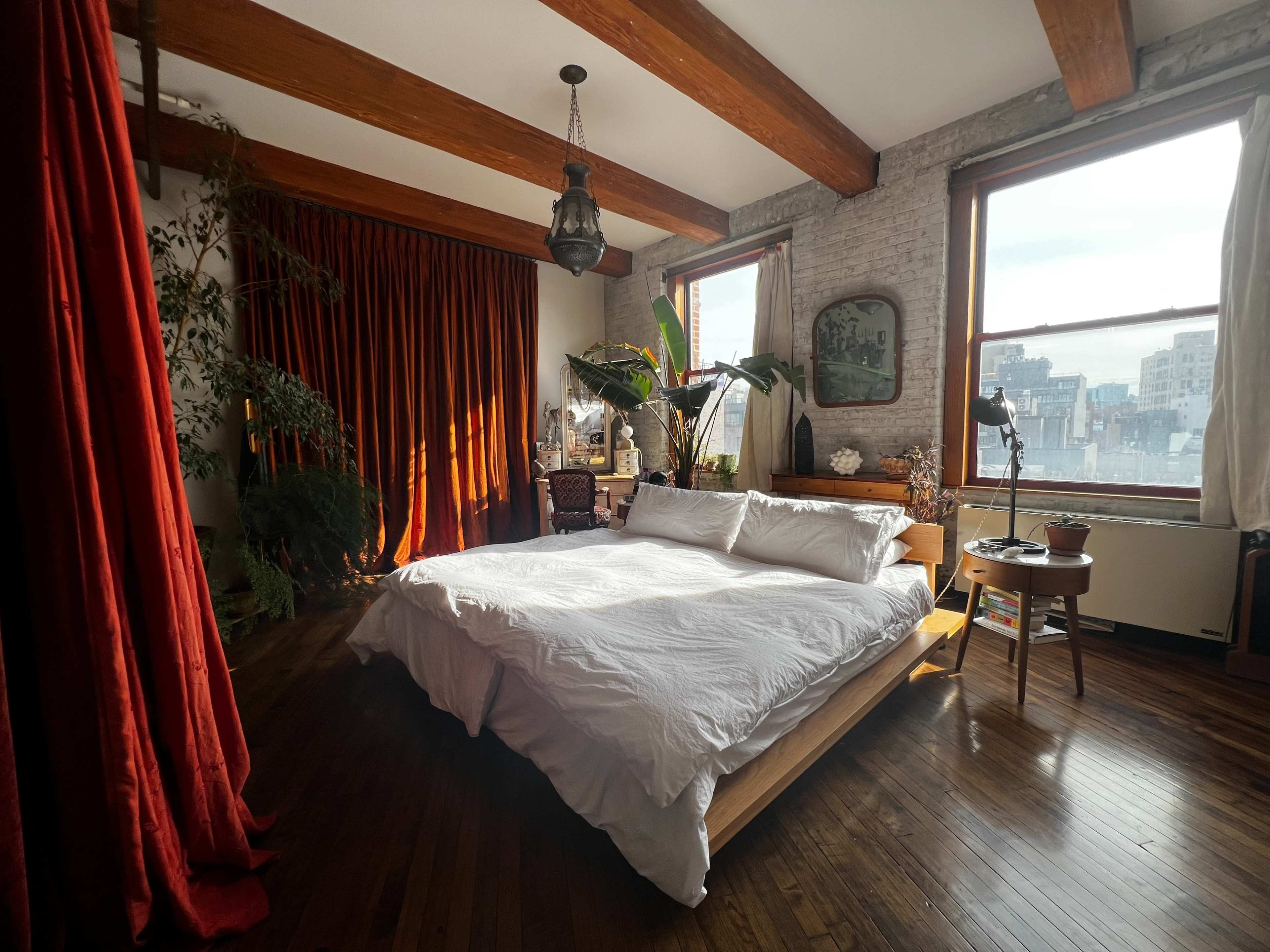 A bright bedroom features a large bed with white bedding, wooden beams on the ceiling, and large windows with red curtains.