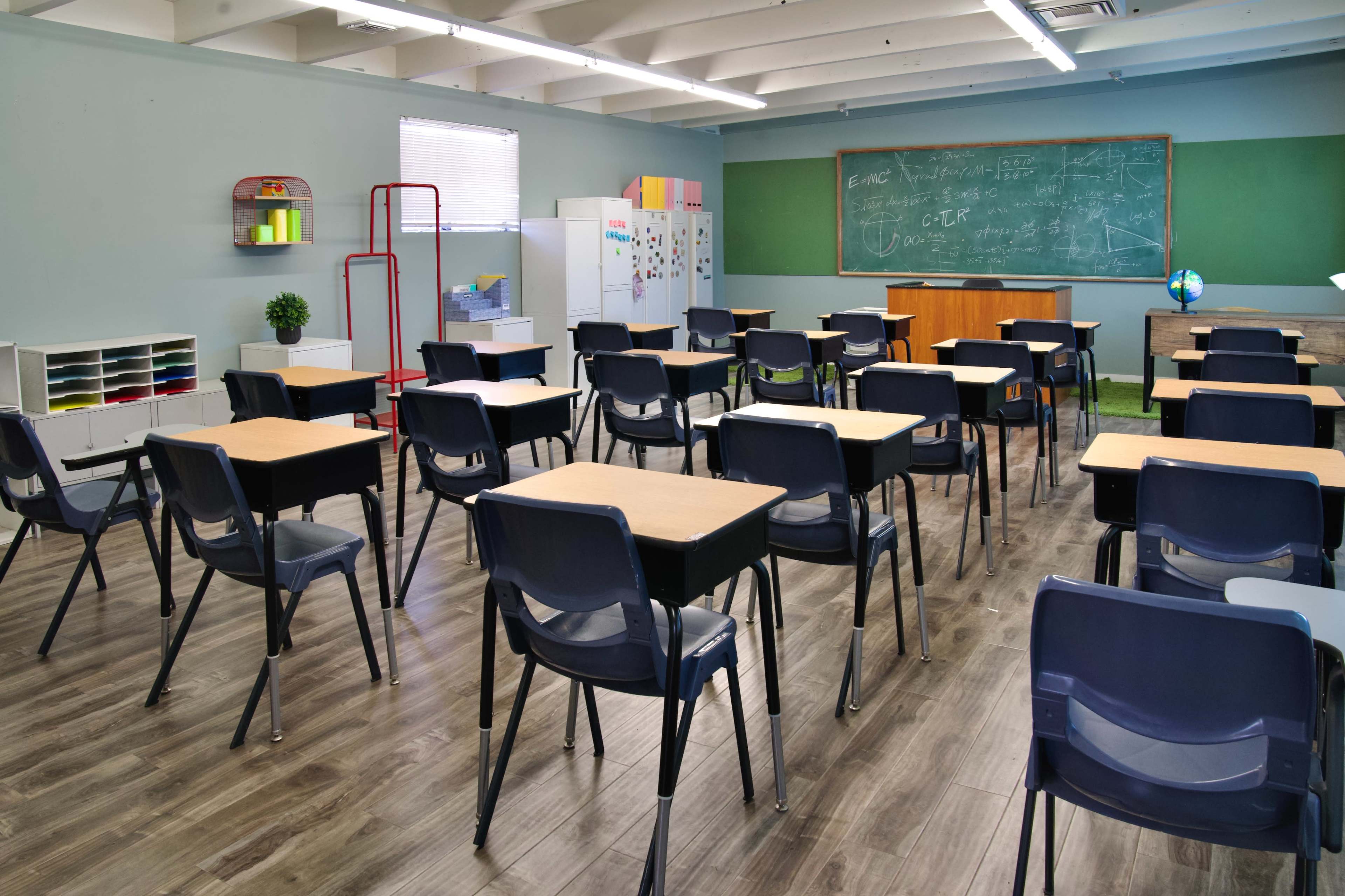 A classroom with empty black chairs and wooden desks arranged in rows, a chalkboard at the front, and various educational materials along the walls.