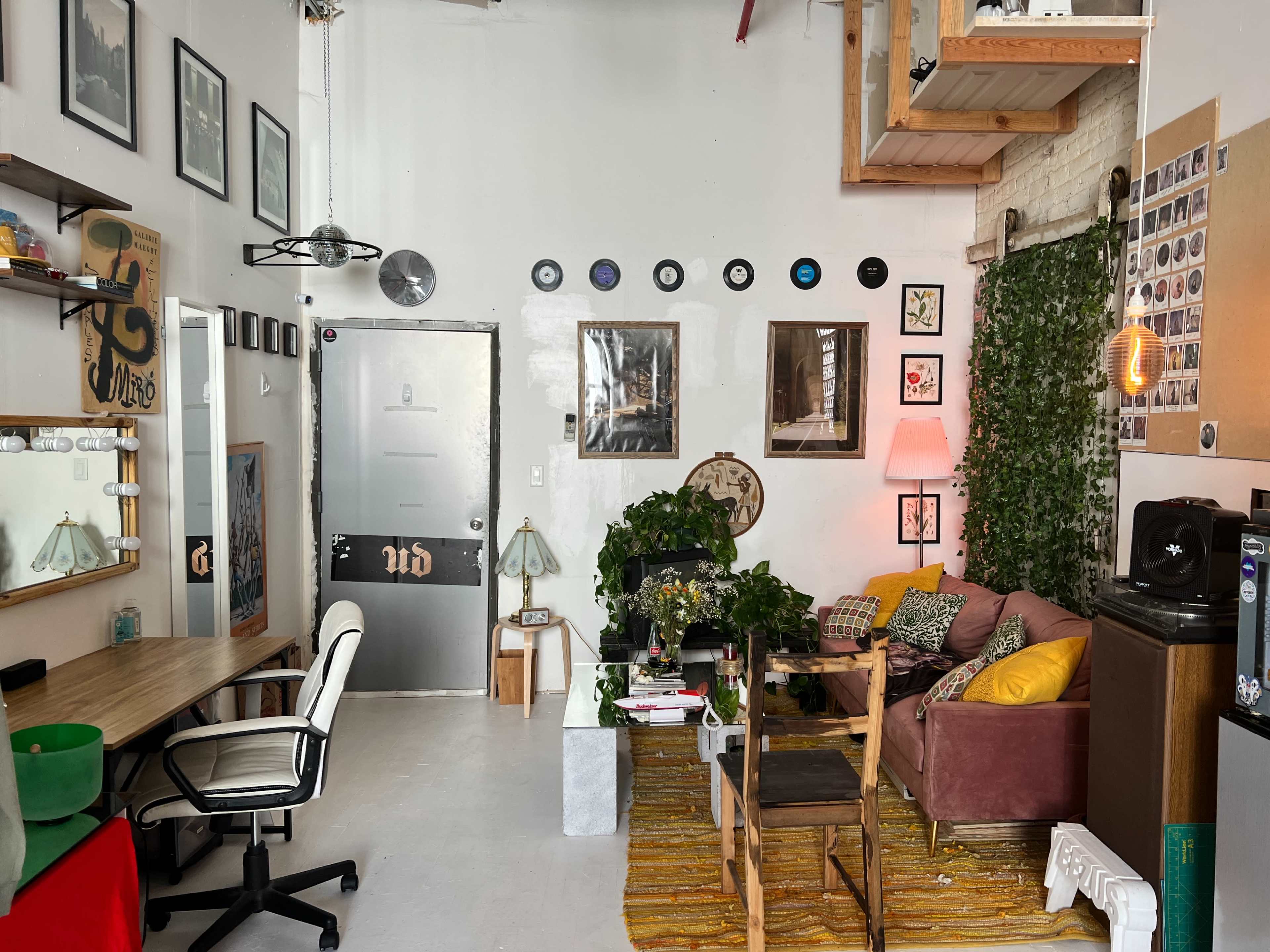 A cozy studio apartment featuring a seating area with a pink sofa, a desk with a chair, and various plants along the walls.