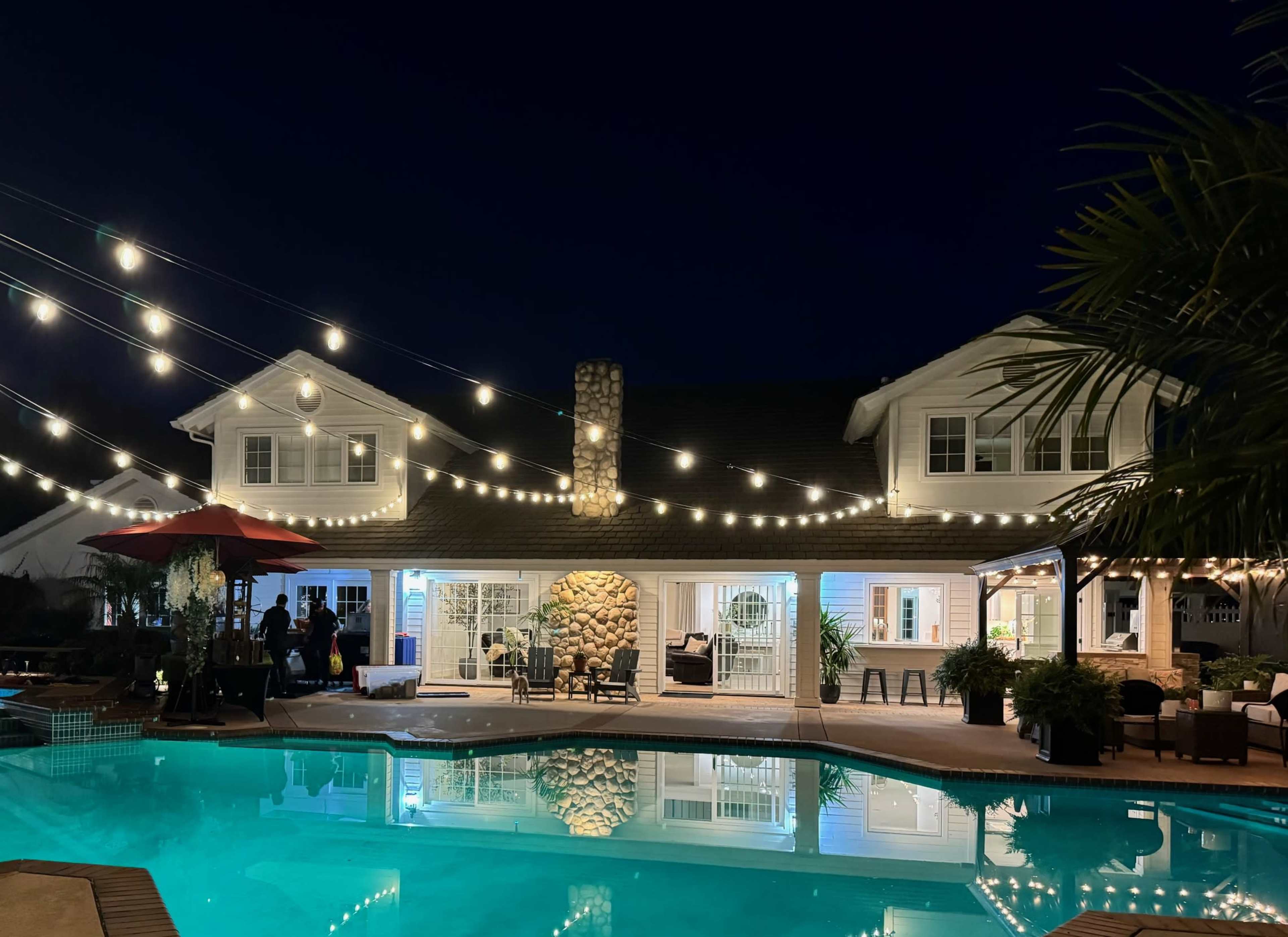 The scene shows a pool area at night with a house featuring stone accents, illuminated string lights overhead, and people gathered around the poolside.