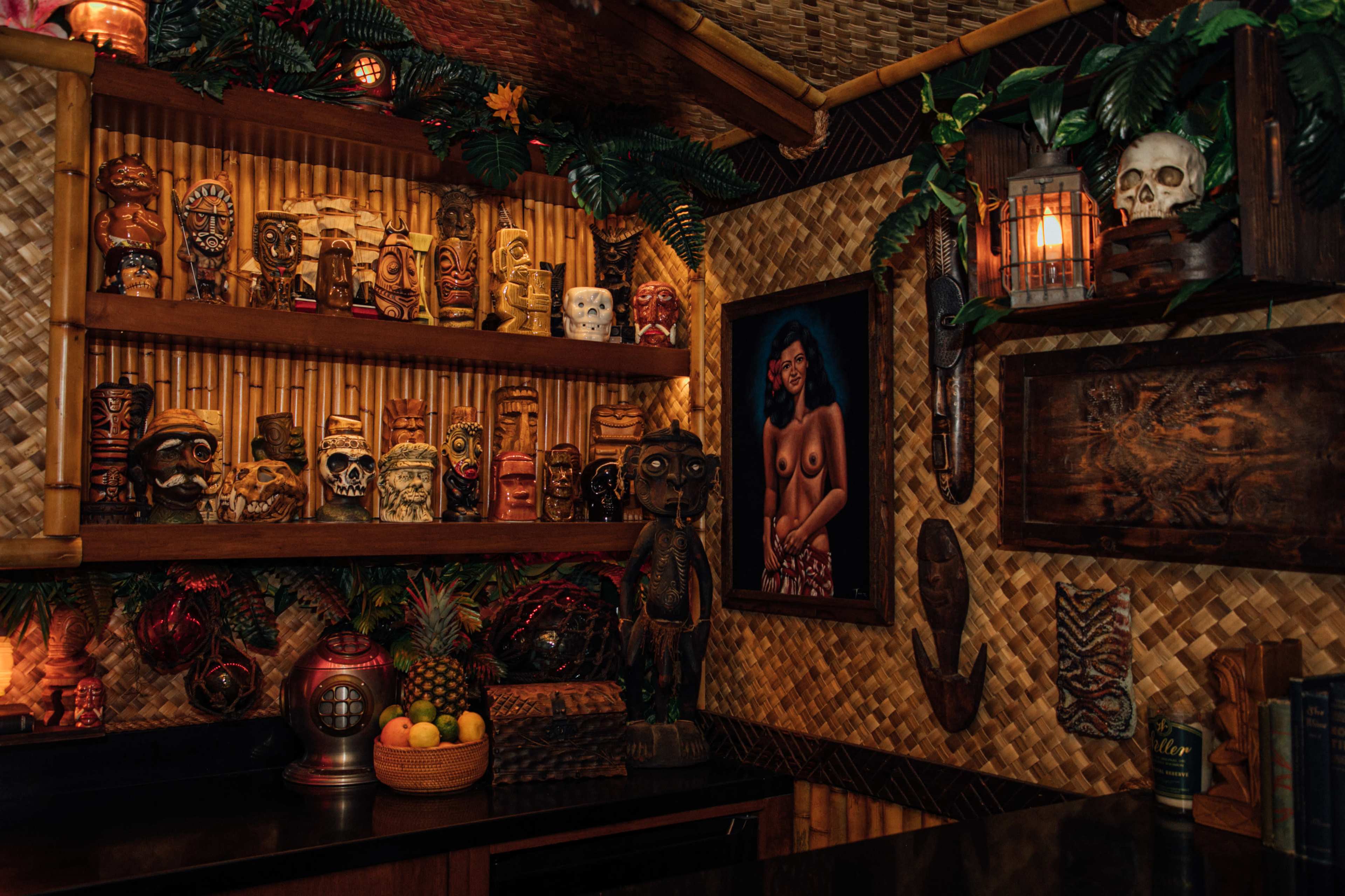 The image shows a dimly lit tiki-themed room featuring a collection of carved wooden masks, colorful mugs, and a framed artwork on a textured wall.