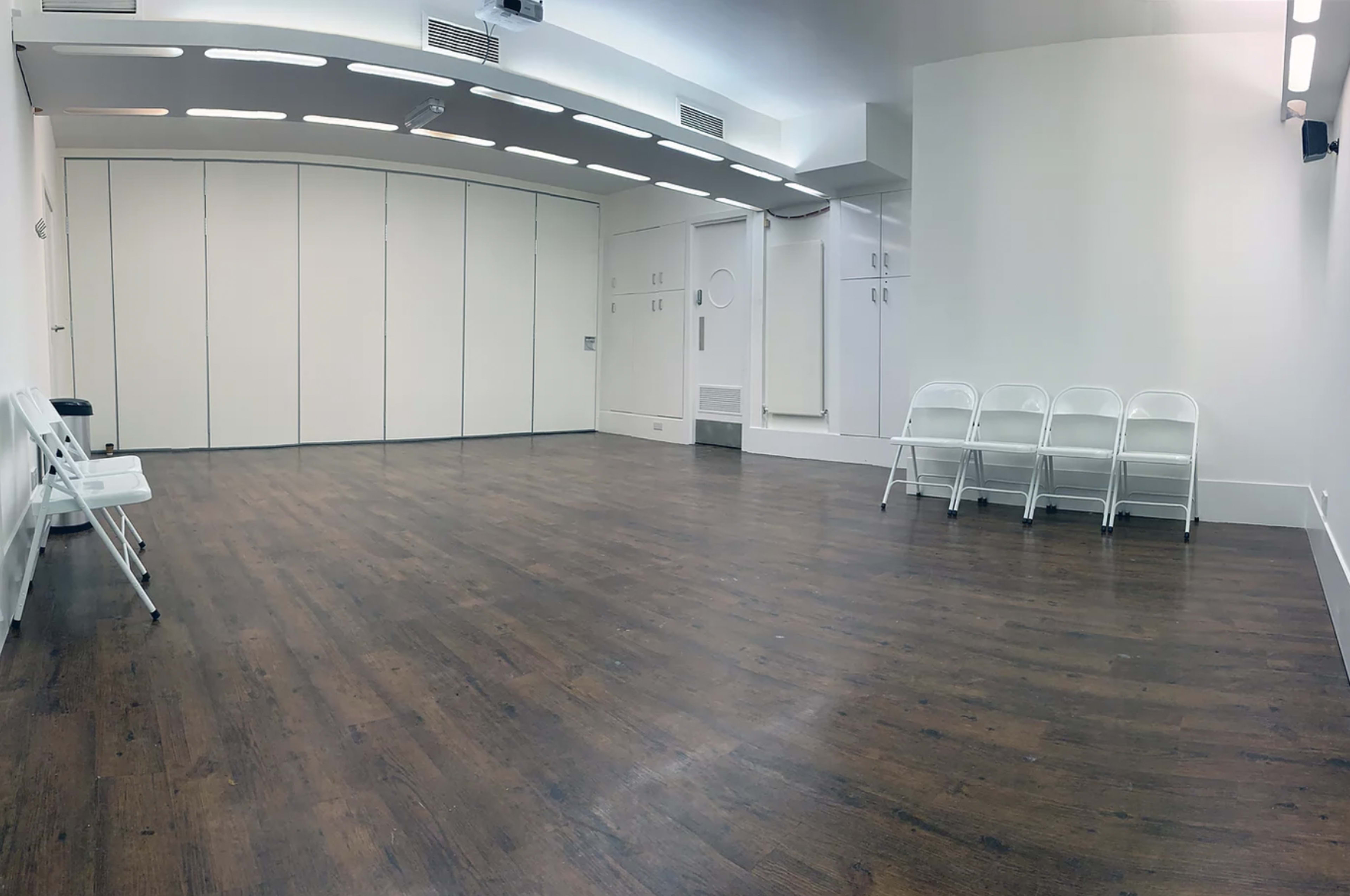 A spacious, empty room with wooden flooring and four white folding chairs arranged against one wall.