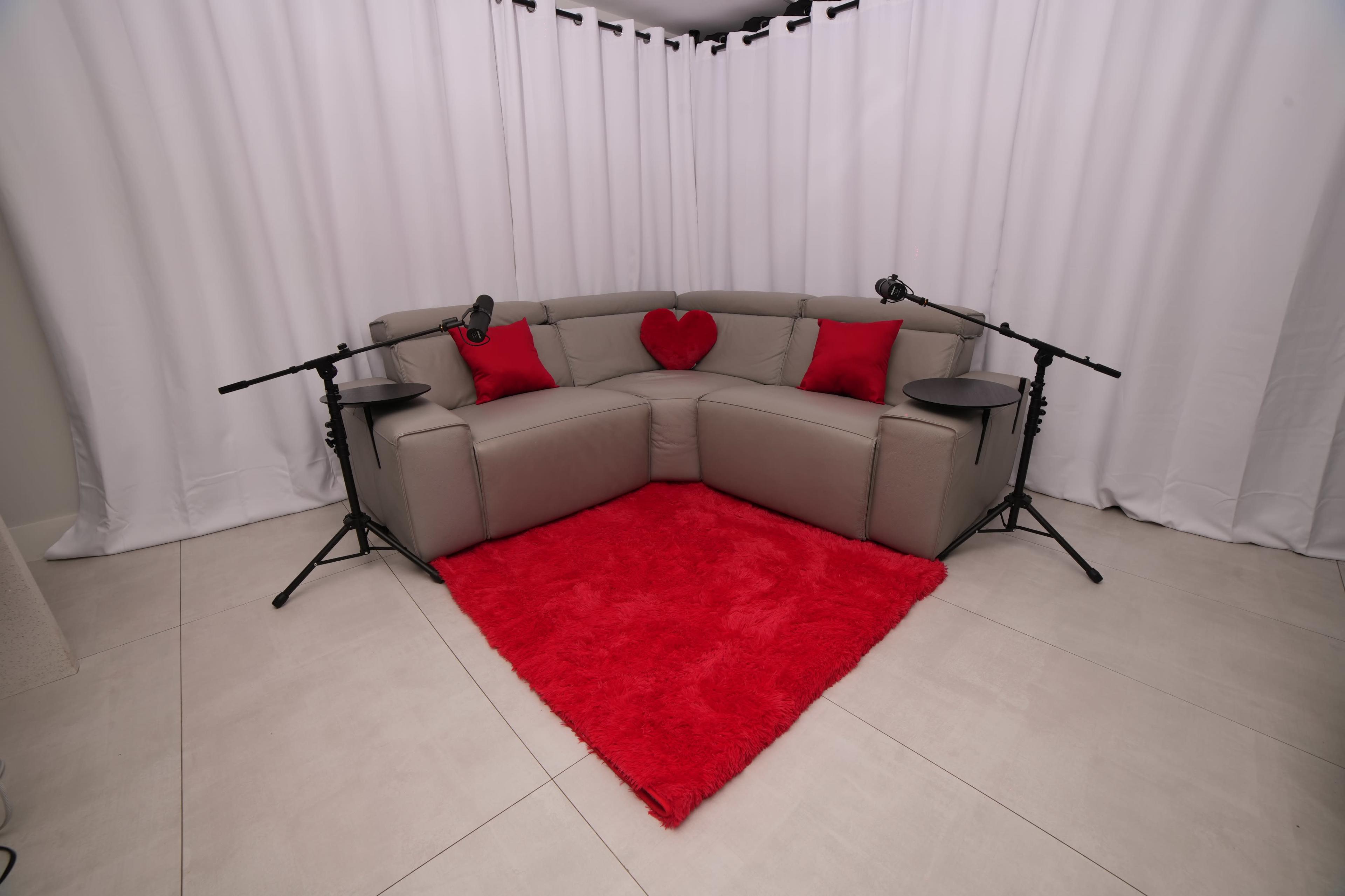 A corner seating arrangement with a grey sectional sofa adorned with red pillows and a heart, accompanied by two black tripods with lights, on a red shag rug, set against white curtains.