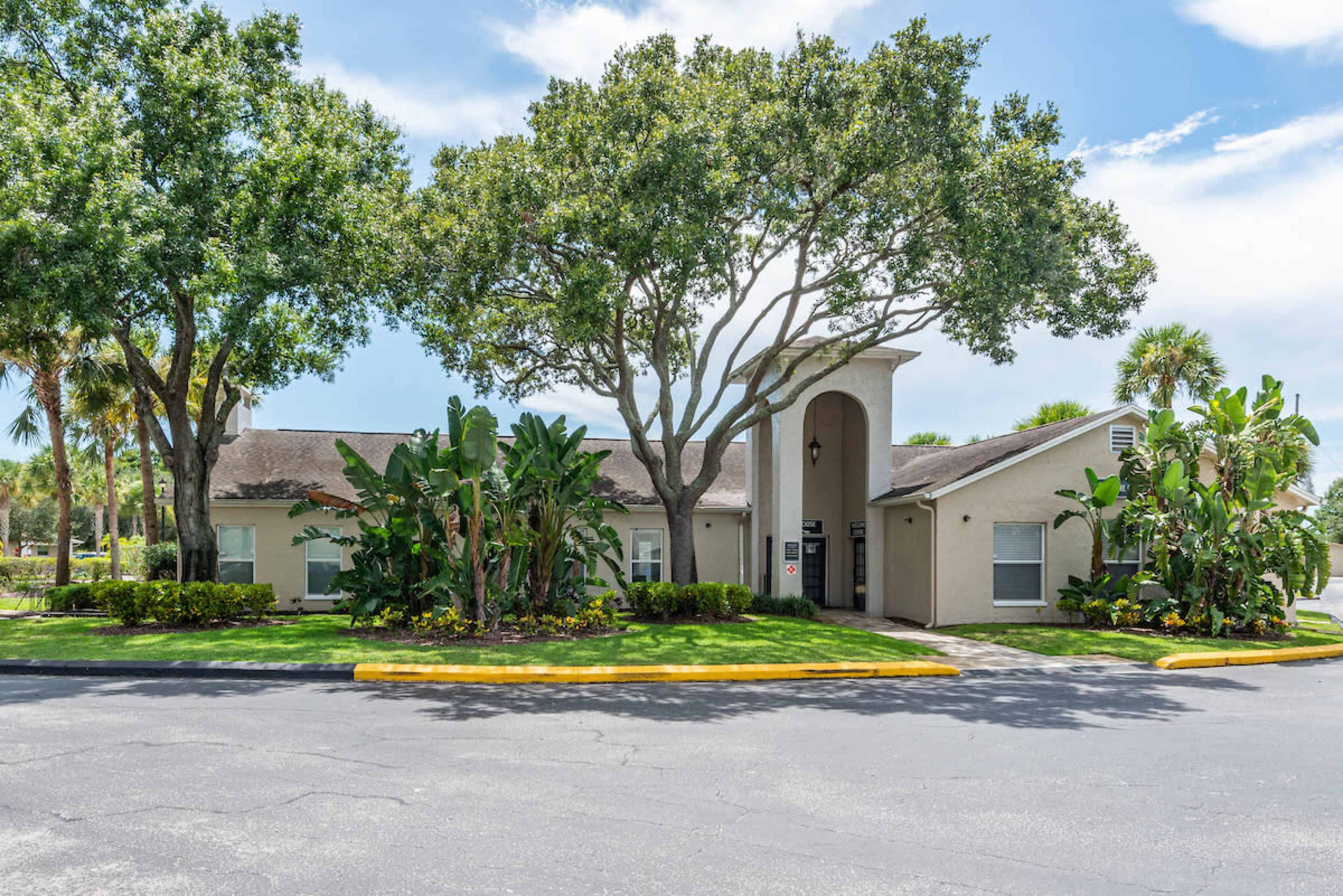 A single-story building surrounded by lush greenery and palm trees, with a paved parking area in front.