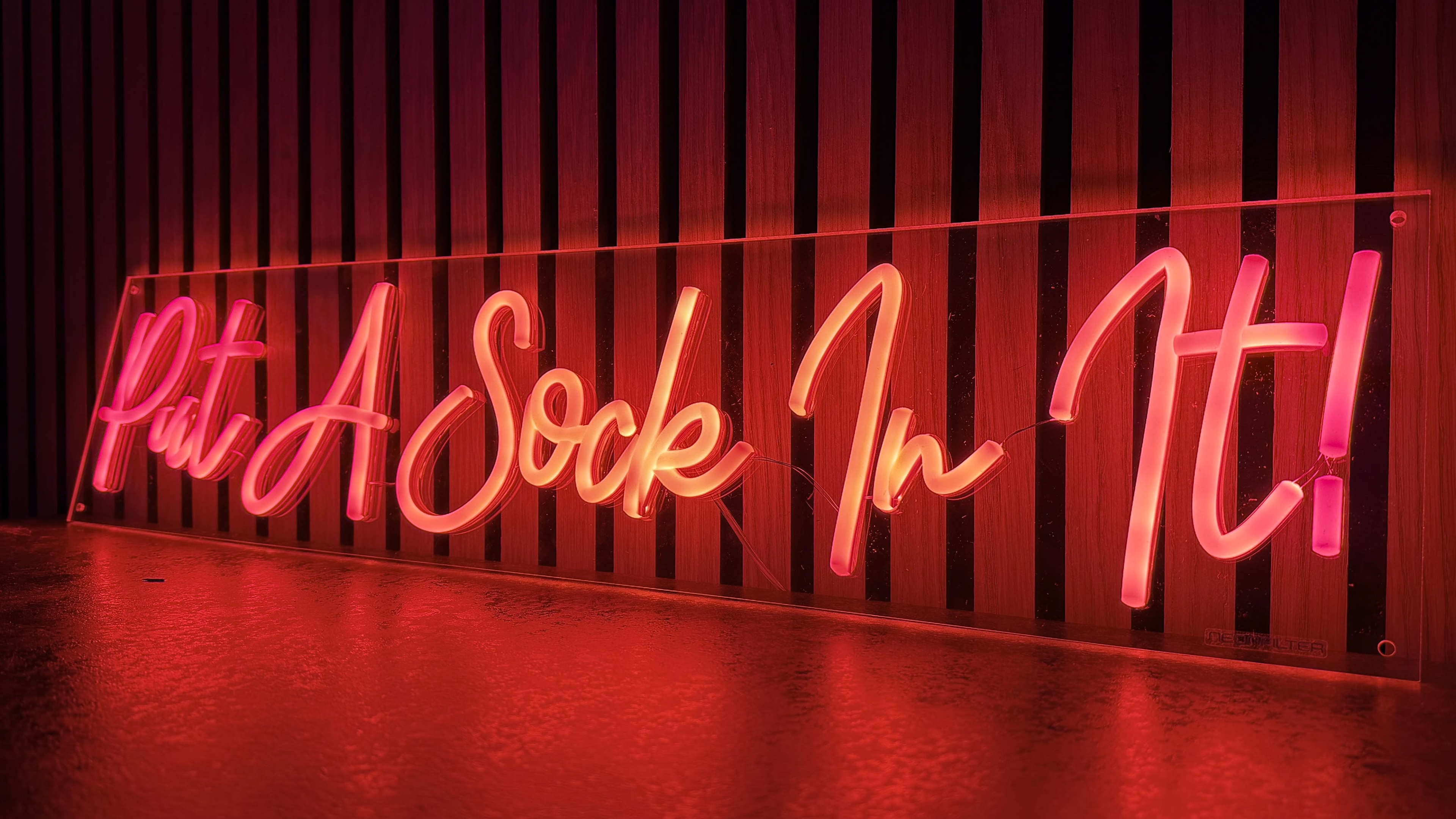 A neon sign displaying the phrase "Put A Sock In It!" is illuminated against a striped background.