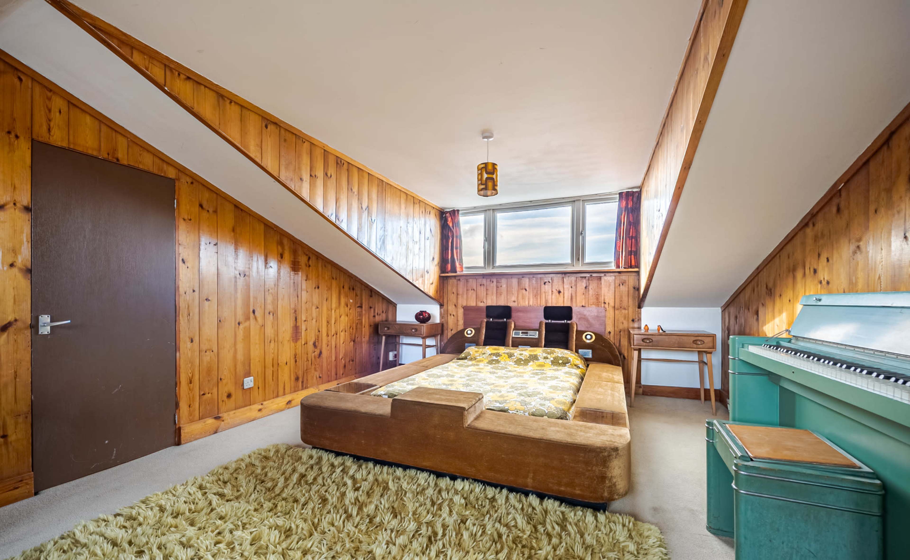 The image shows a cozy room with wooden paneled walls, a bed with a patterned blanket, a piano, and a small desk by a window.