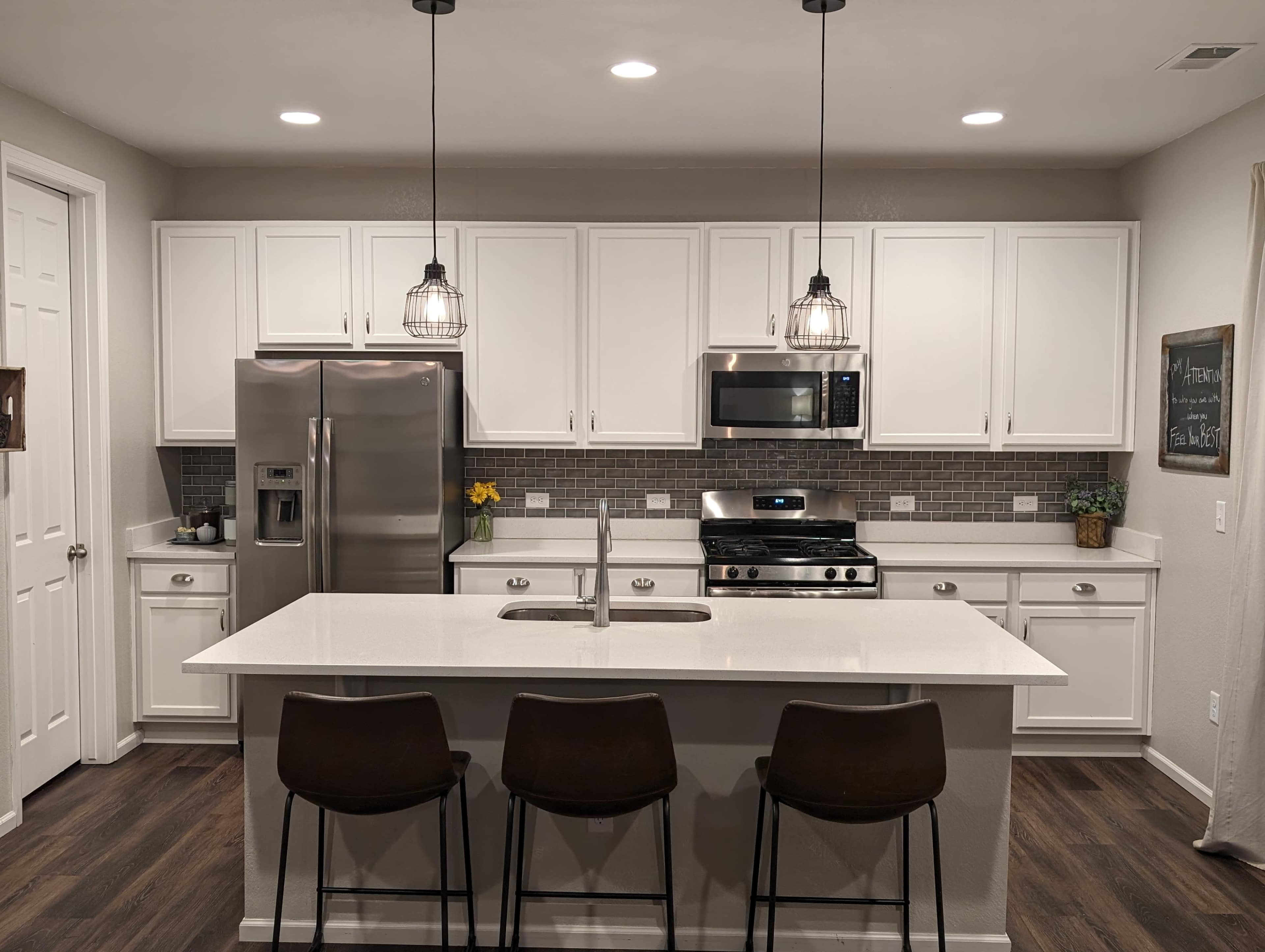 A modern kitchen featuring white cabinets, stainless steel appliances, a central island with three bar stools, and pendant lighting.