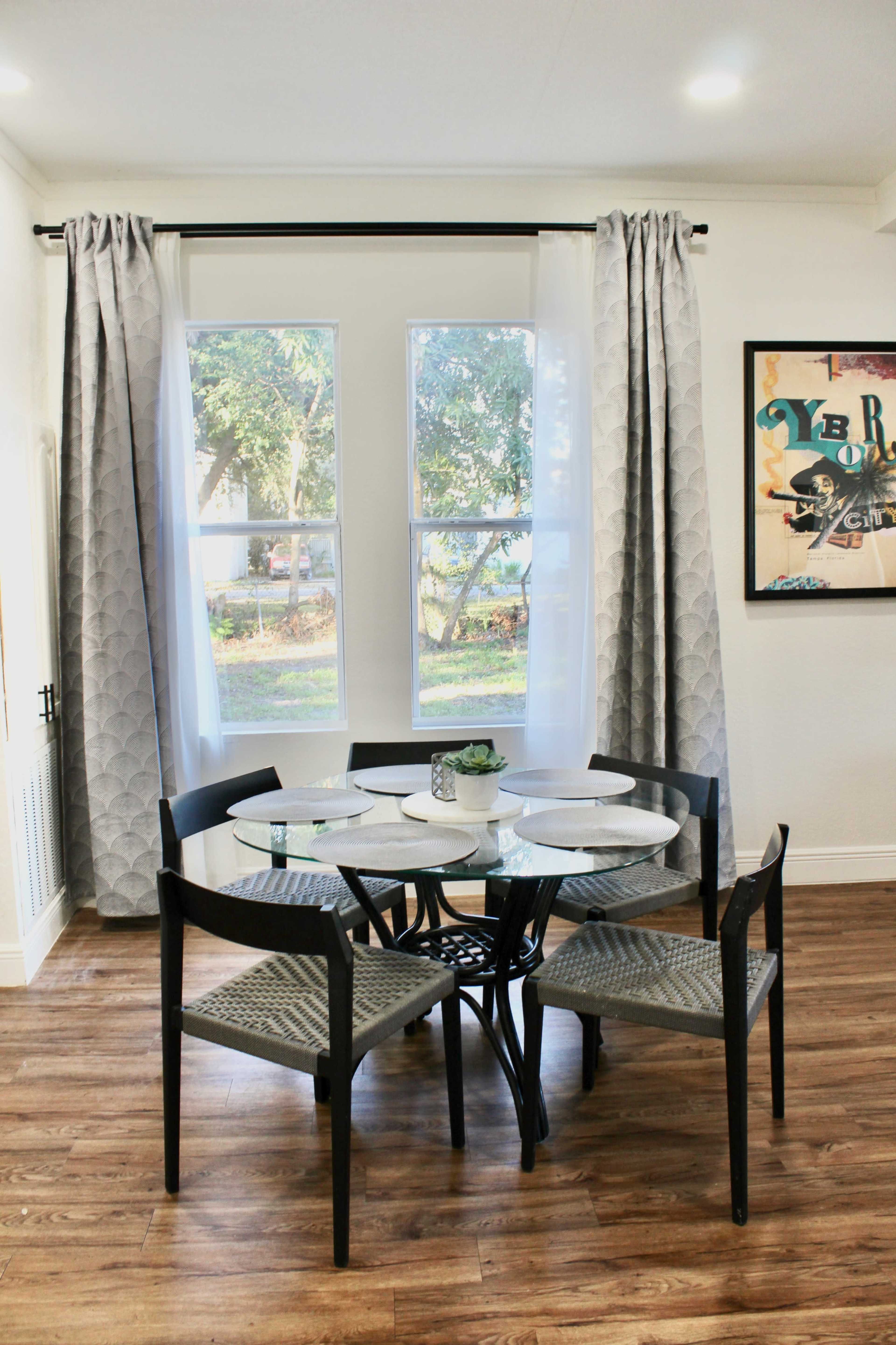 A round dining table with four chairs is positioned under two large windows draped with gray curtains in a well-lit room.