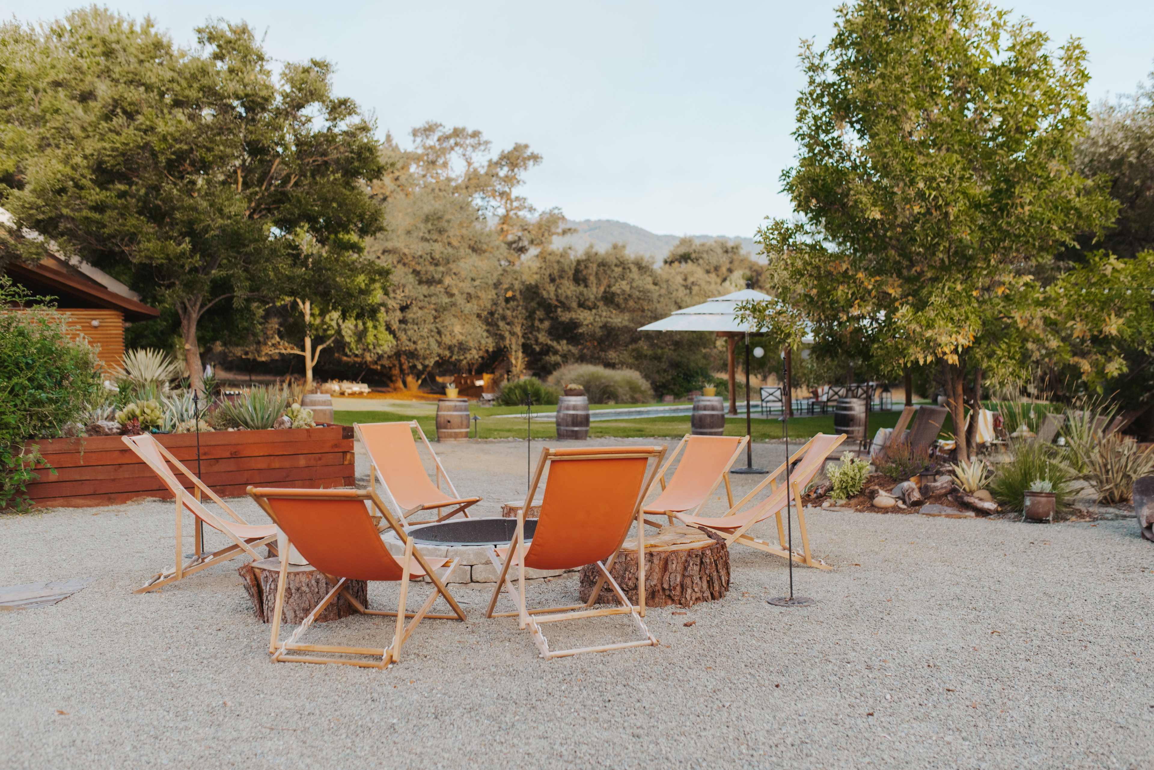 A circular arrangement of orange chairs surrounds a fire pit made from a tree stump in a gravel outdoor area.