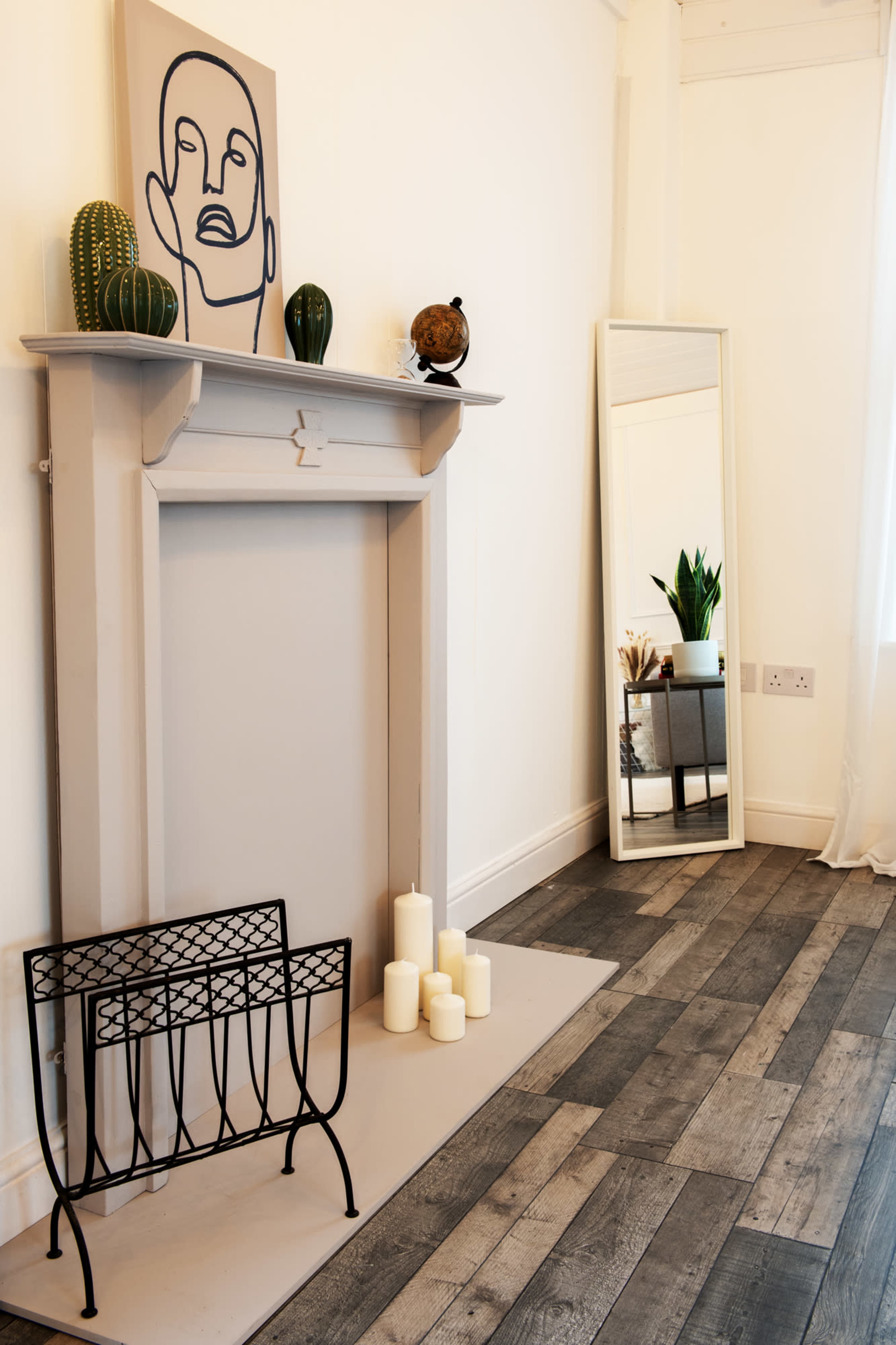 A minimalist interior featuring a decorative fireplace with a wall-mounted artwork, candles, a potted plant, and a standing mirror beside it.