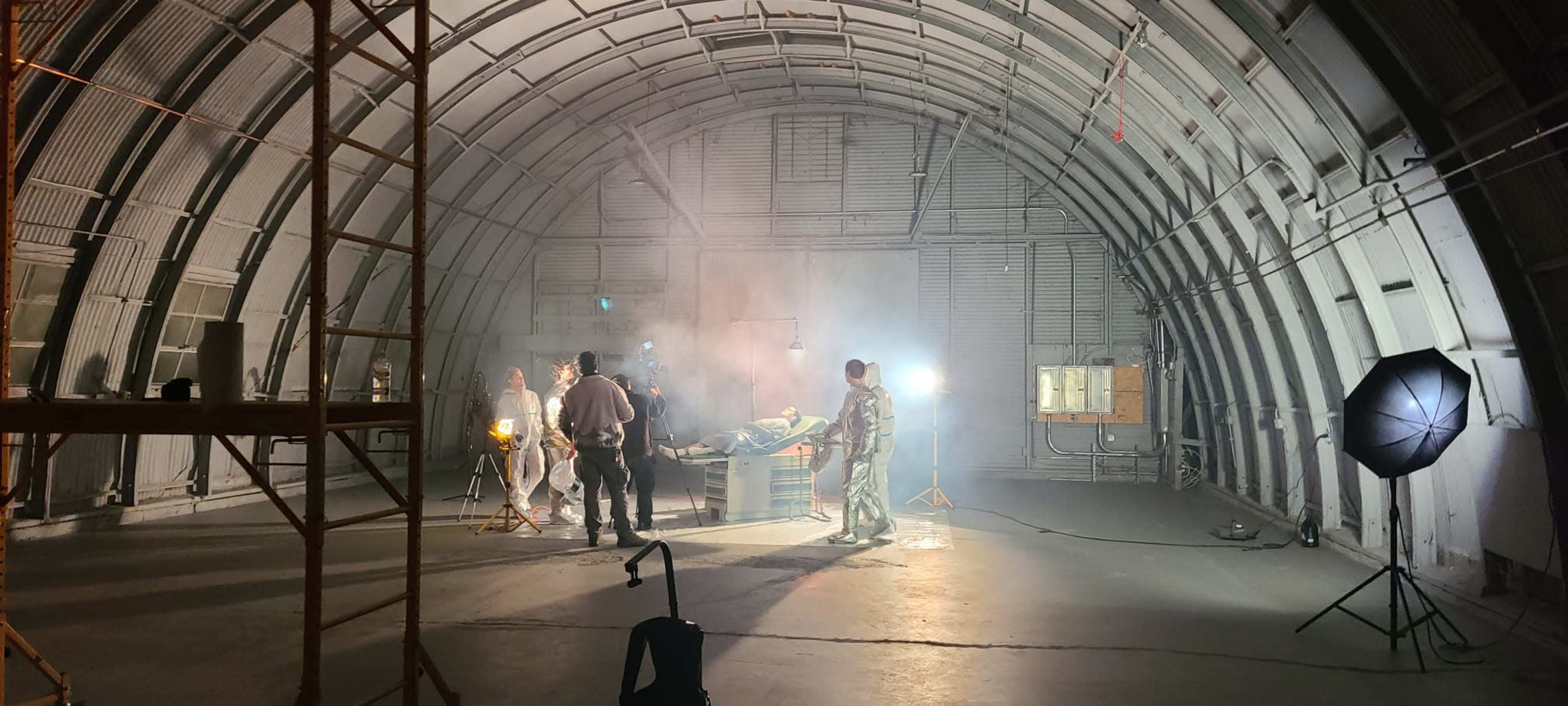 A group of people, including military personnel, are working inside a large, industrial-style building that has scaffolding, lighting equipment, and smoke effects.