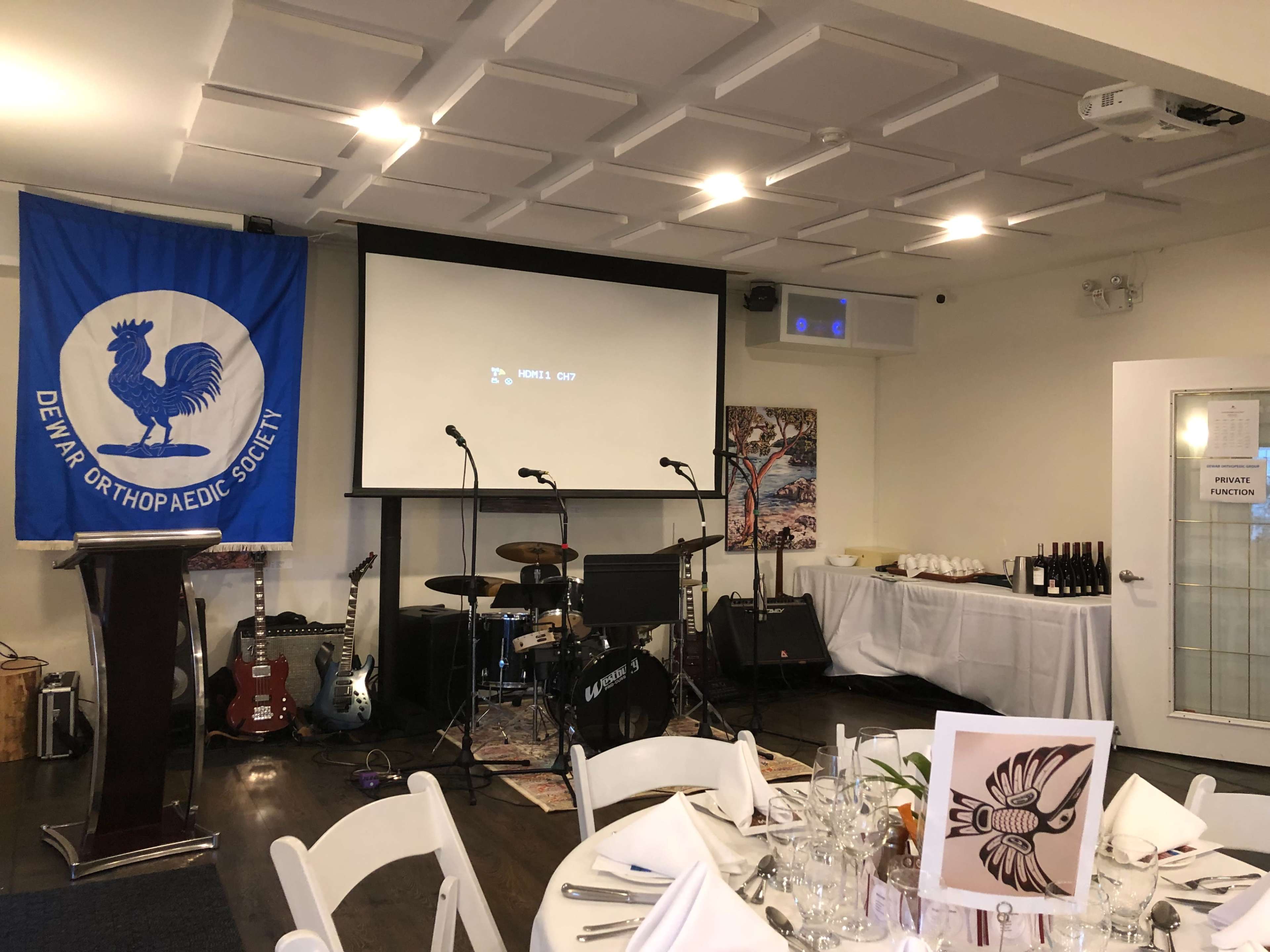 A room set up for a private function, featuring a stage with musical equipment, a large screen, and a table with food and drinks.