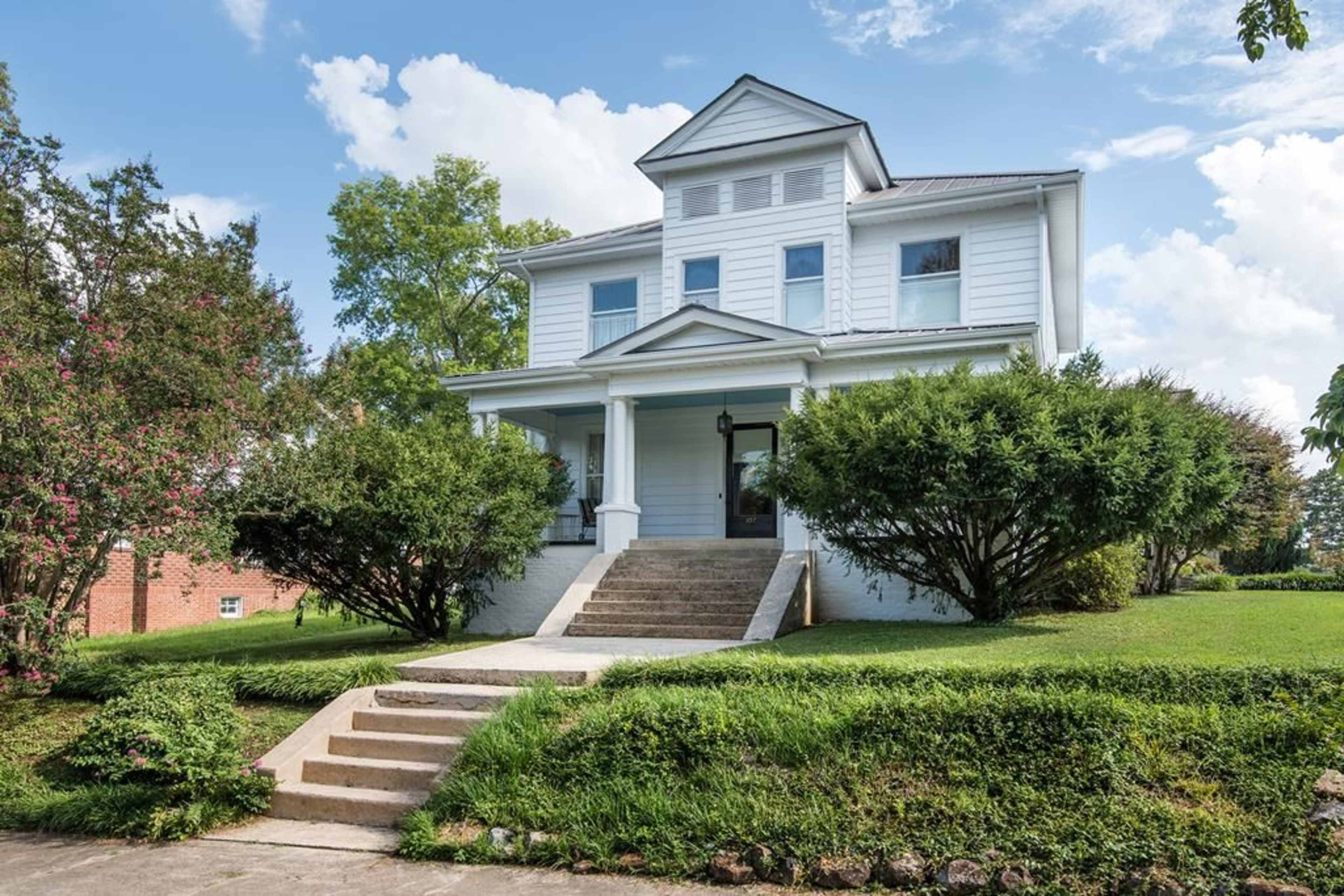 A two-story white house with a front porch, situated on a landscaped yard with shrubs and steps leading to the entrance.