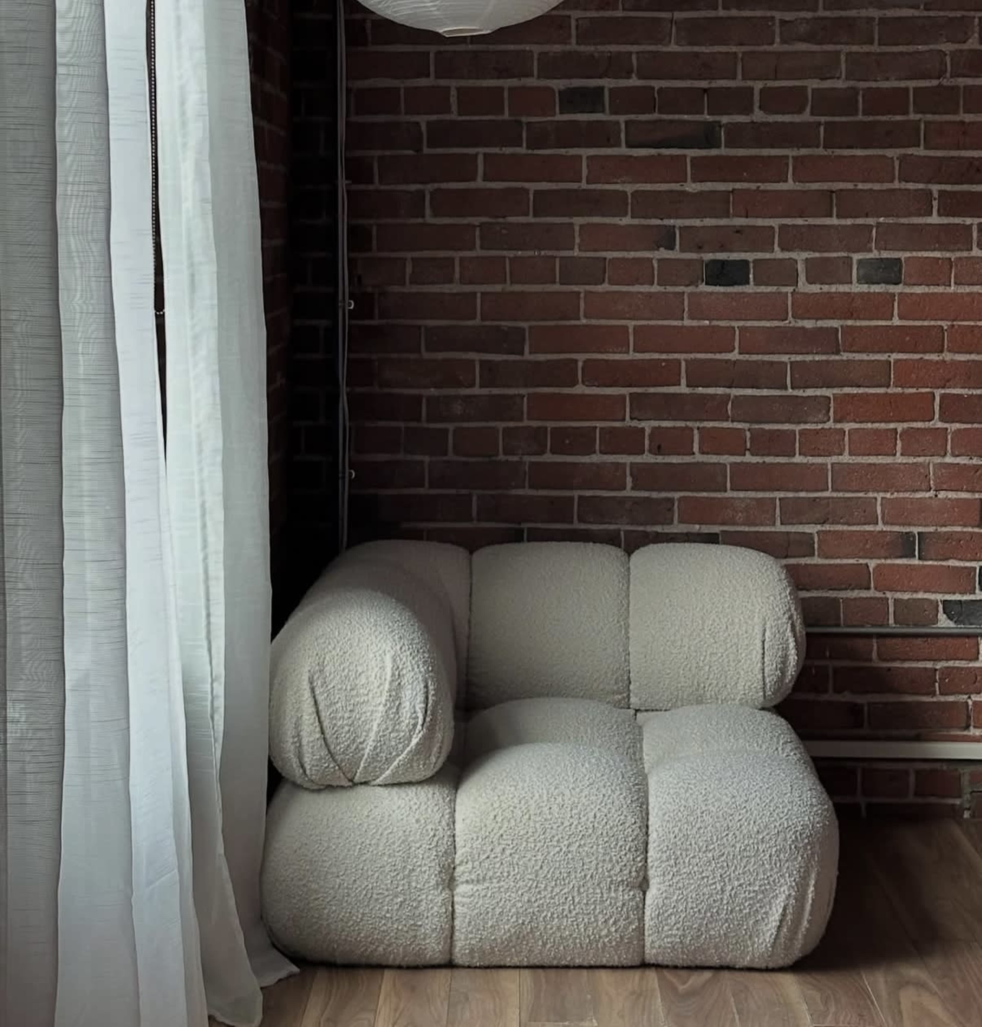 A light-colored, textured two-seater couch is positioned against a brick wall, with sheer white curtains partially covering the window.