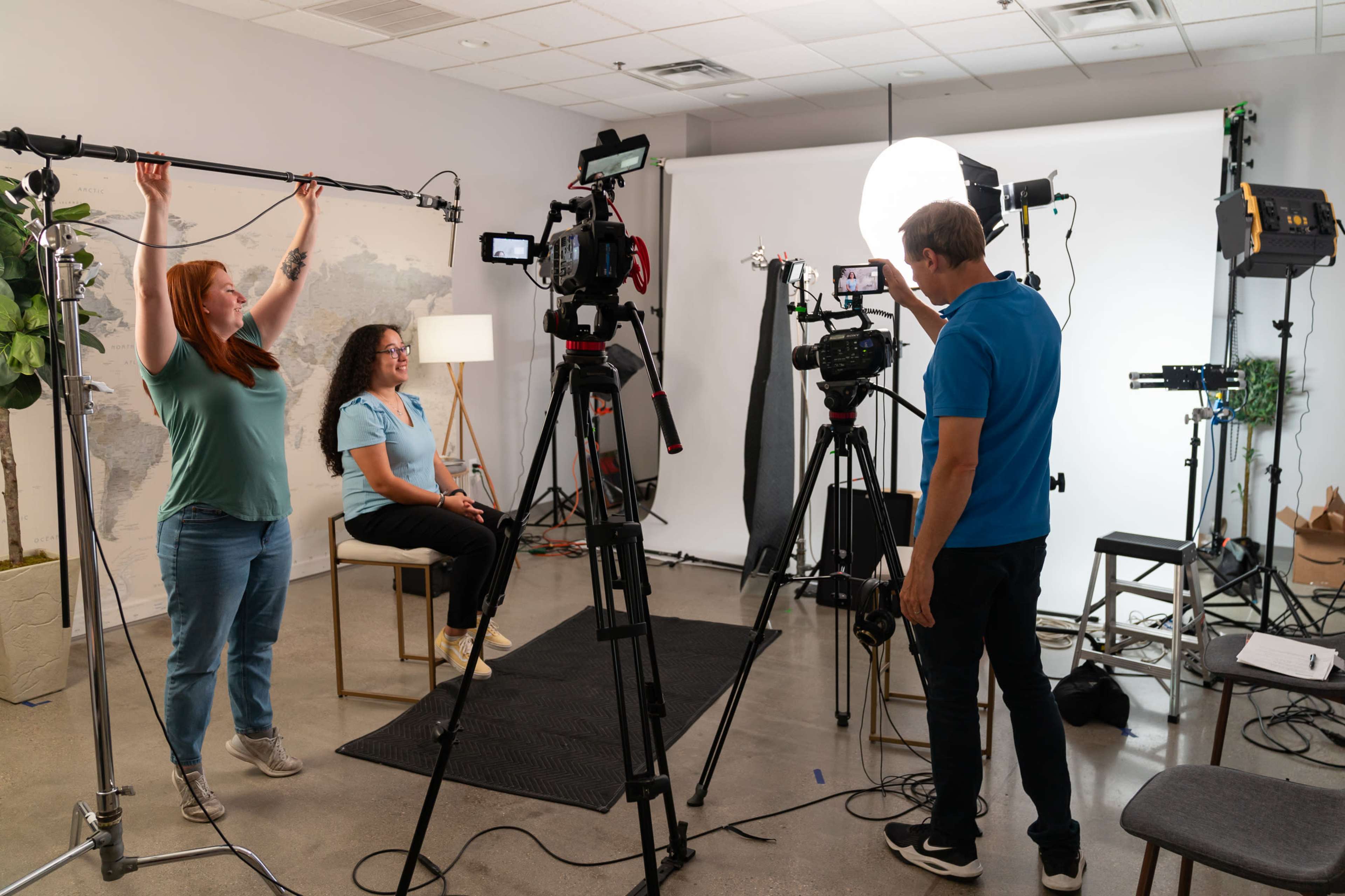 A video production team sets up a shoot, with one person adjusting a microphone while another directs a camera towards a seated subject among various studio lights and equipment.