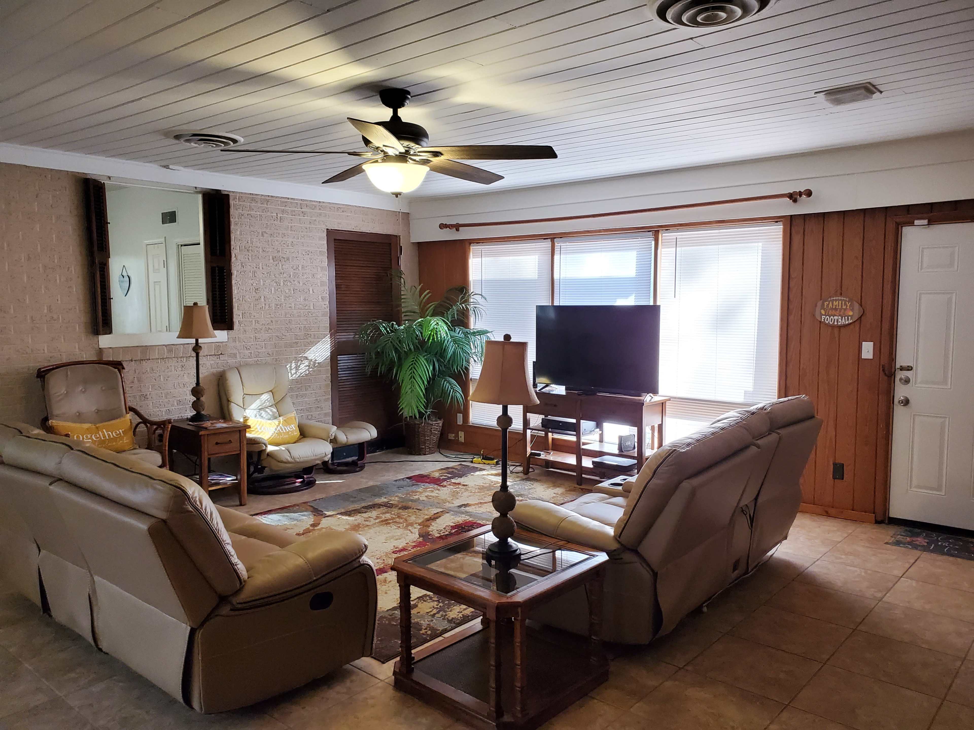 A living room with two leather sofas facing a coffee table, a television in the corner, and large windows allowing natural light.