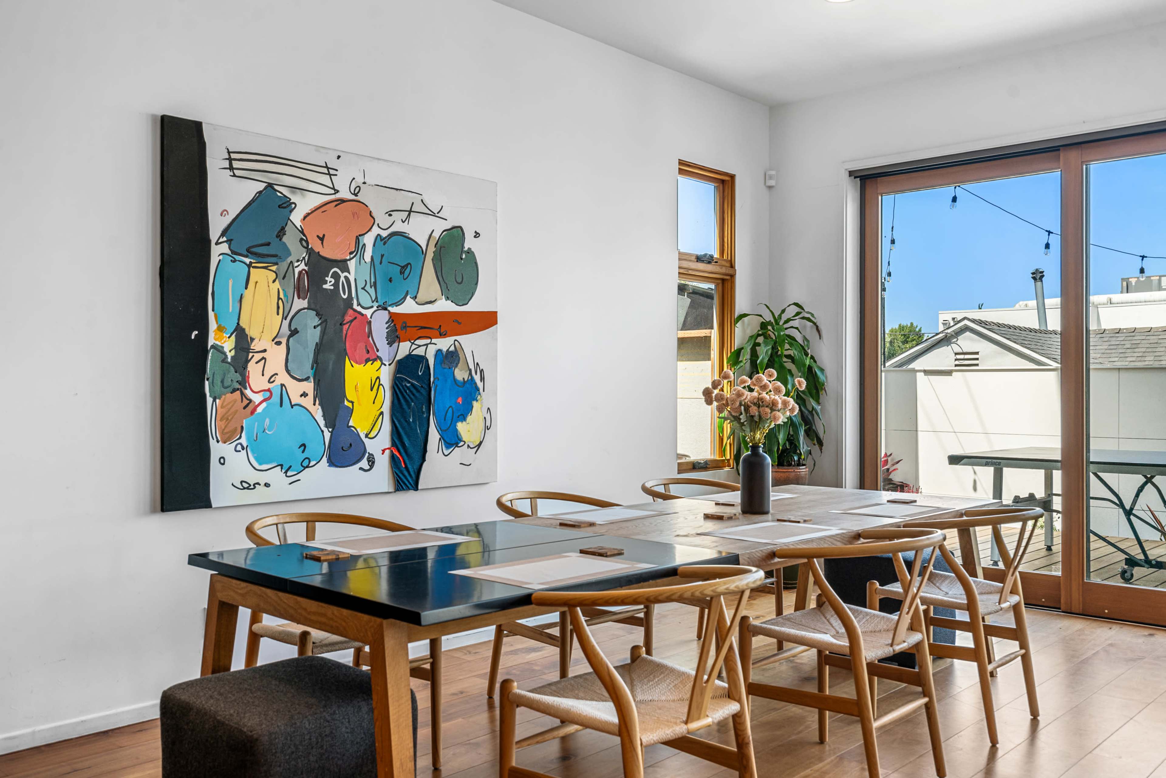 A dining area features a large table surrounded by wooden chairs, with a colorful abstract painting on the wall and sliding glass doors that open to an outdoor view.