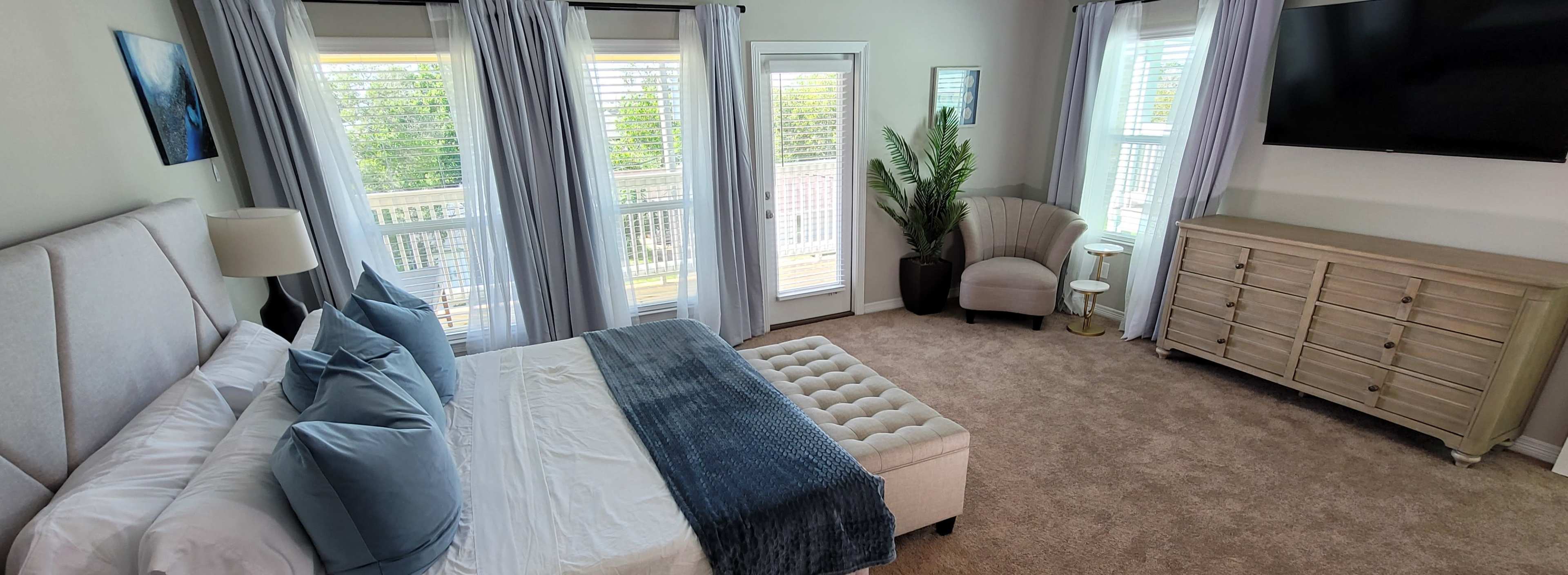 A spacious bedroom features a large bed with blue accents, a sliding glass door leading to a balcony, and ample natural light from two windows.